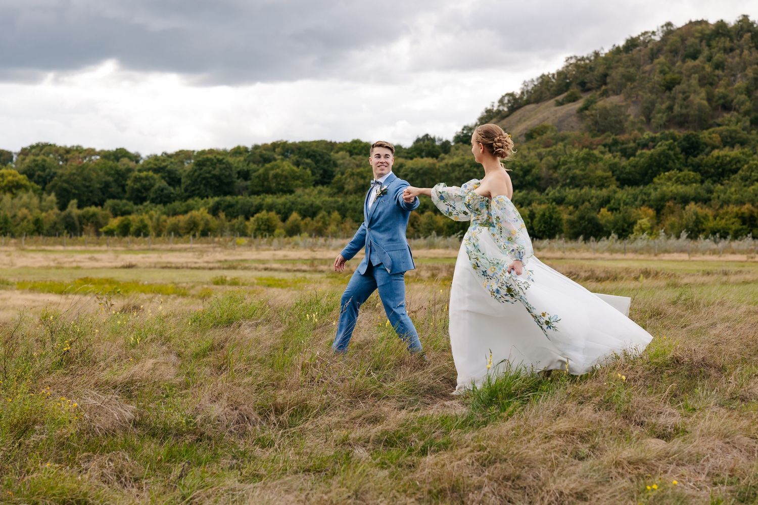 Bride and groom holding hands walking through a scenic field with a green hill in the background.