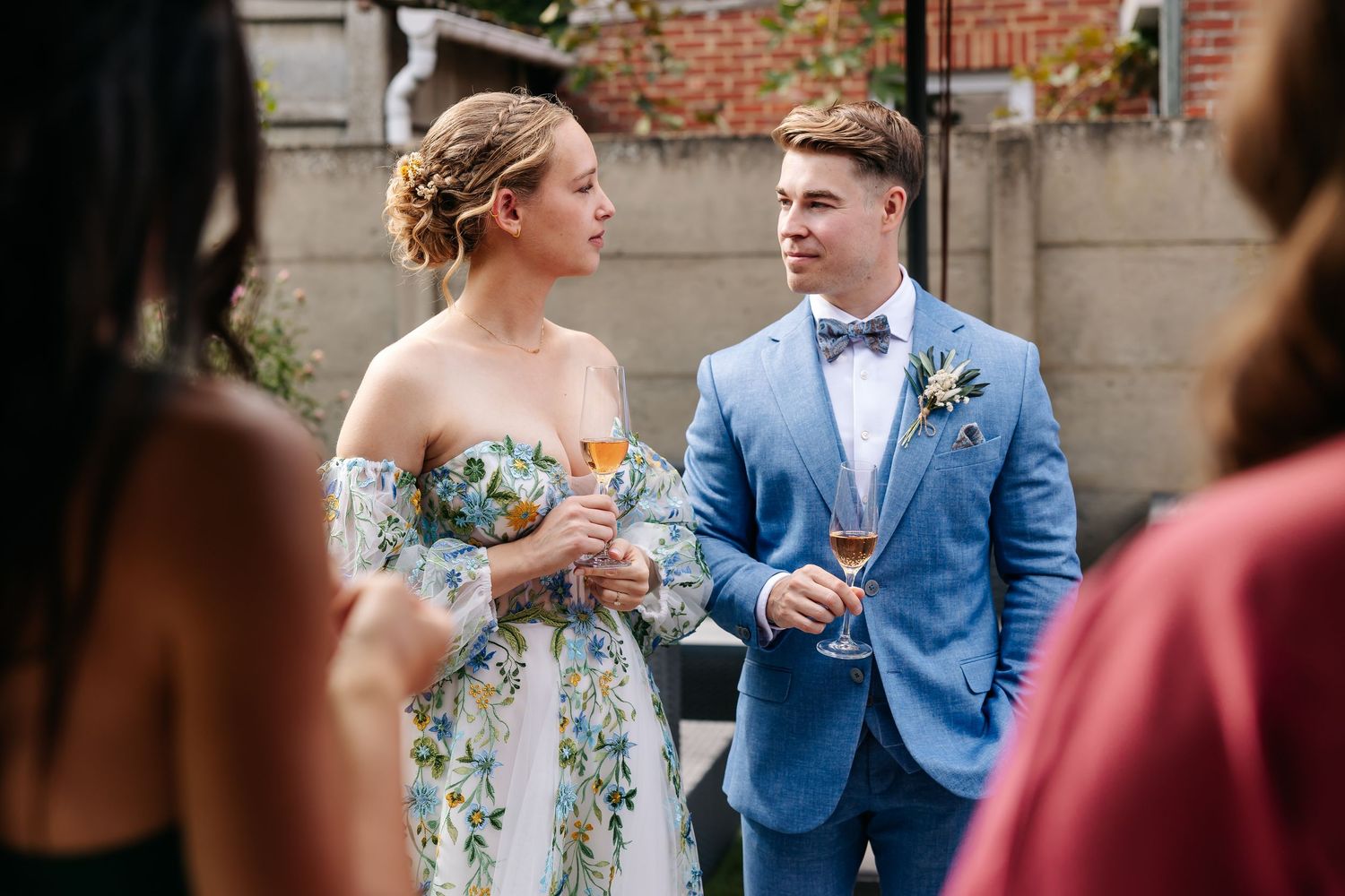 Bride in floral dress and groom in blue suit chatting with wedding guests.