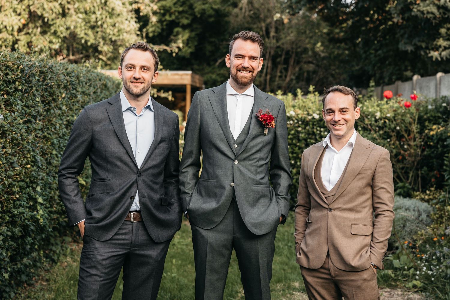 Three men in suits posing in garden, groom in gray three-piece suit with red boutonniere at wedding.