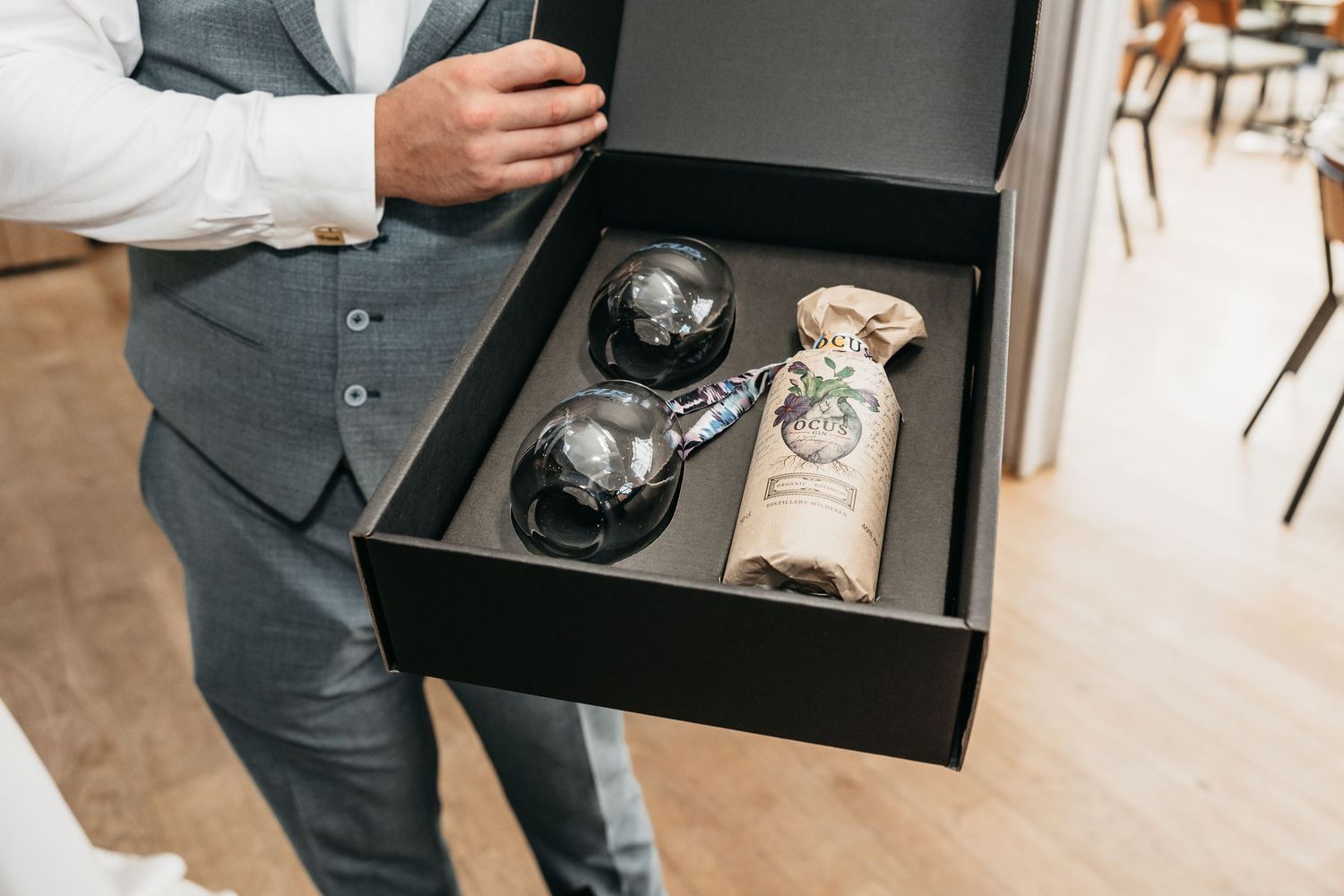 Groom in grey vest presenting an elegant black gift box containing wine bottle and decorative black glasses.