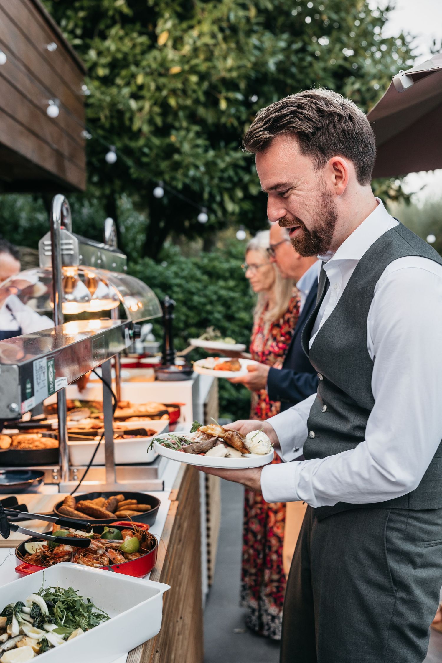 Well-dressed man selecting food from an outdoor catering buffet station at an evening event.