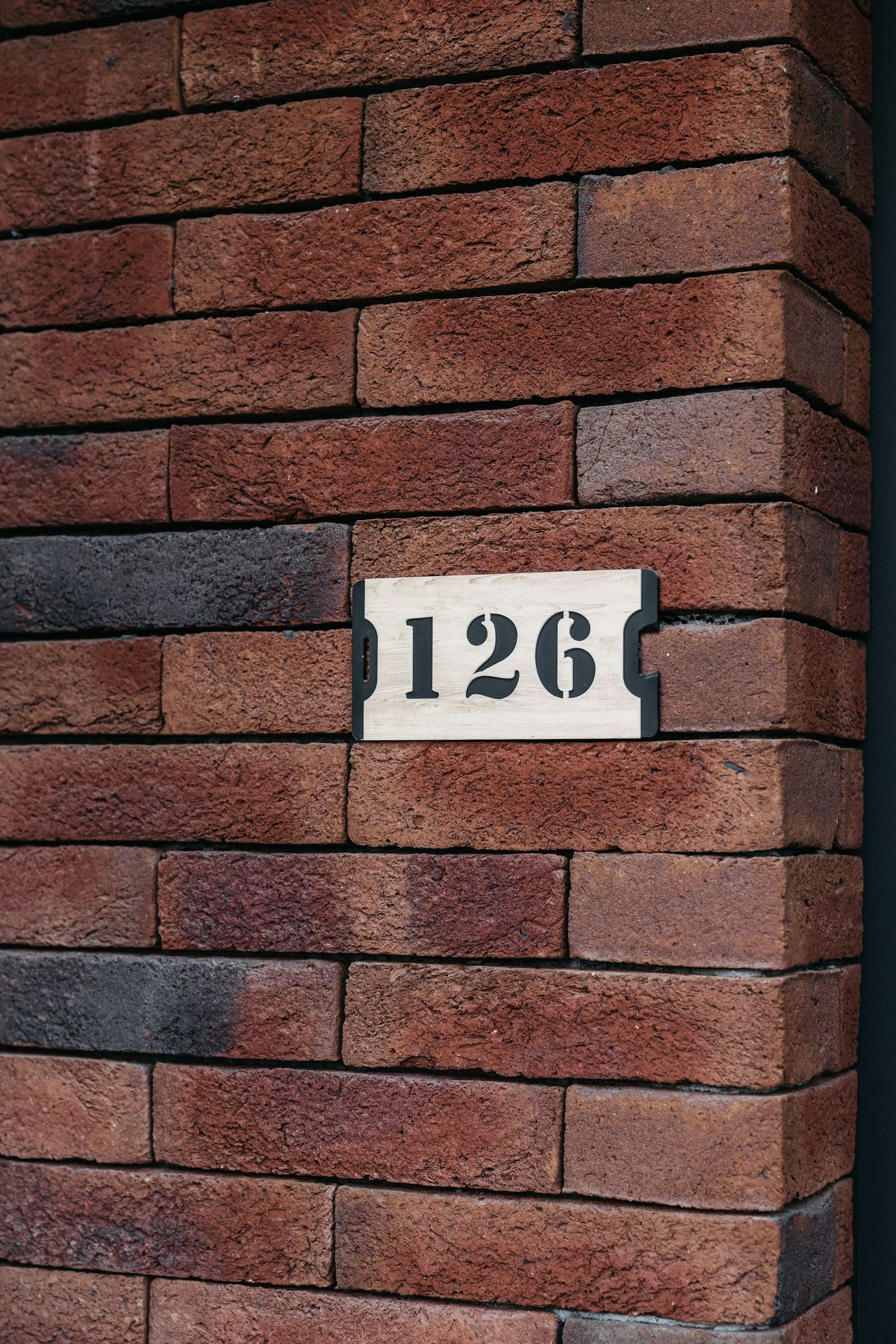 Close-up of a white number 126 address plaque mounted on a red brick wall corner.