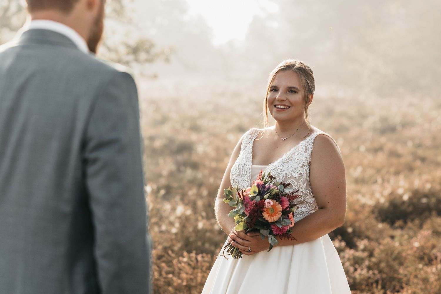 Smiling bride in lace wedding gown holds colorful bouquet while facing her groom in a misty outdoor setting.