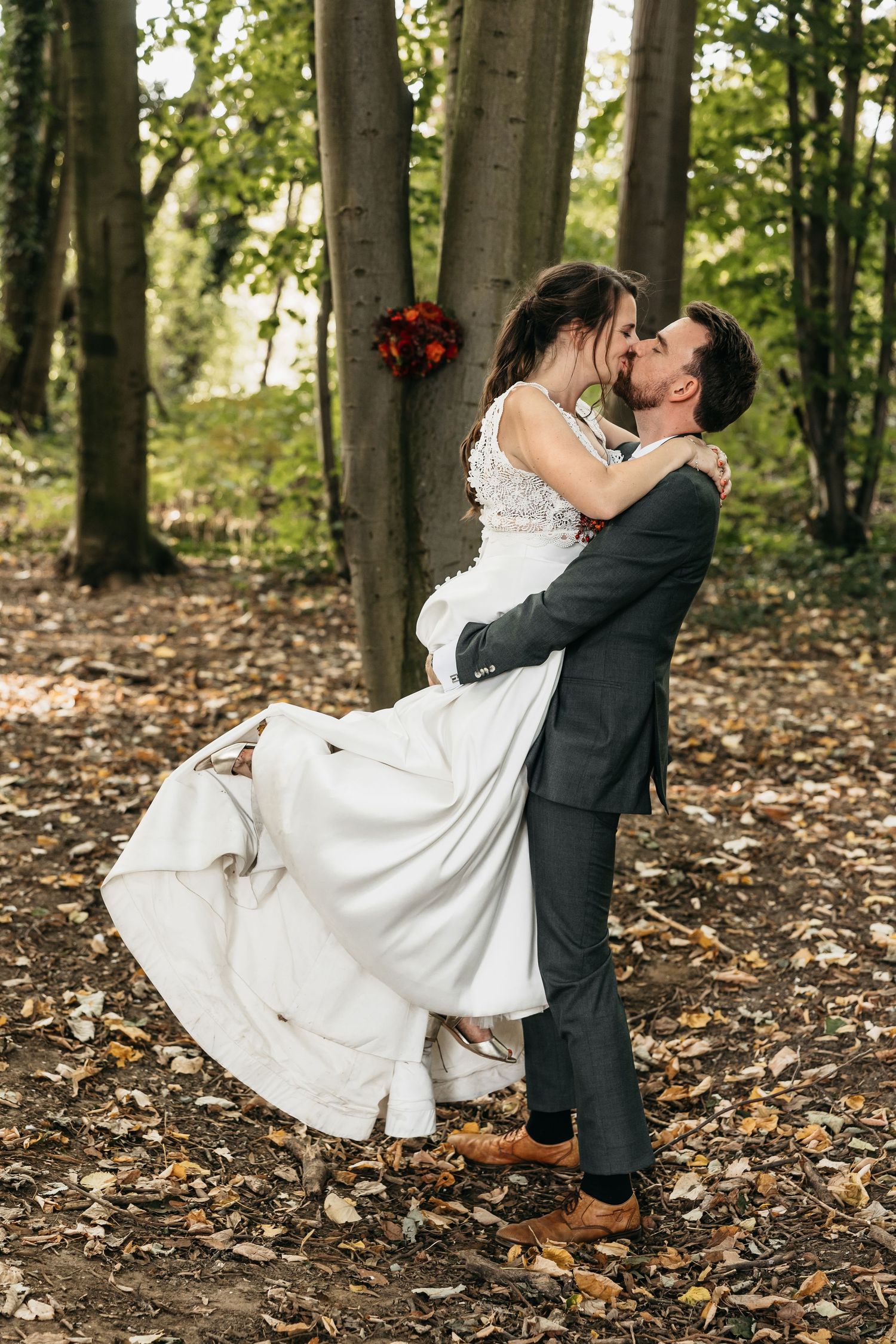Groom lifting bride in white dress for a romantic kiss in a sunlit autumn forest.