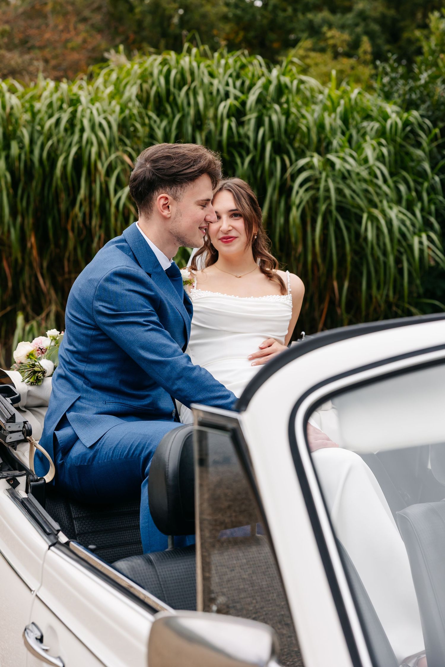 Bride and groom sharing a romantic moment seated together in a classic white vintage convertible wedding car.