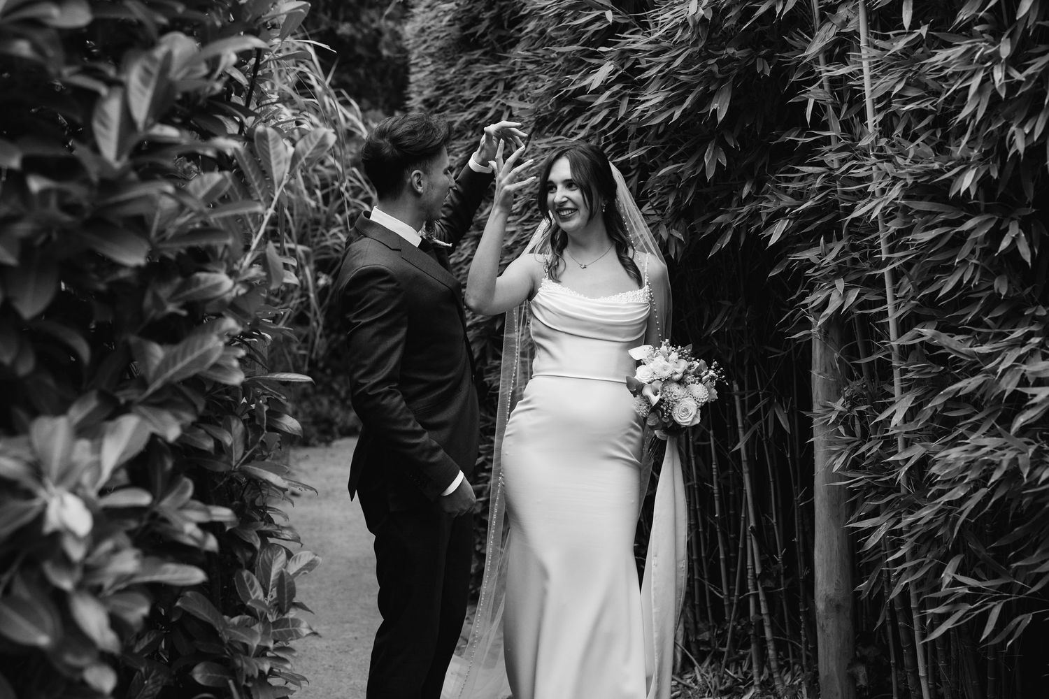 Black and white photo of groom lifting brides veil as she laughs in a bamboo garden.