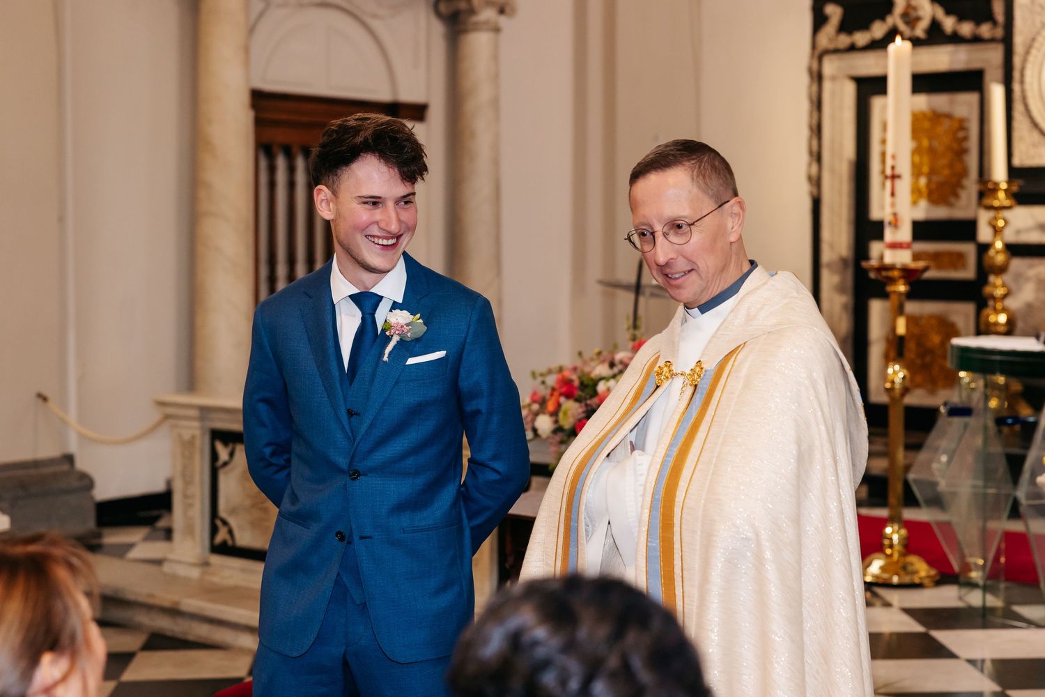 Smiling groom in blue suit with boutonniere standing beside priest in white vestments inside a church.