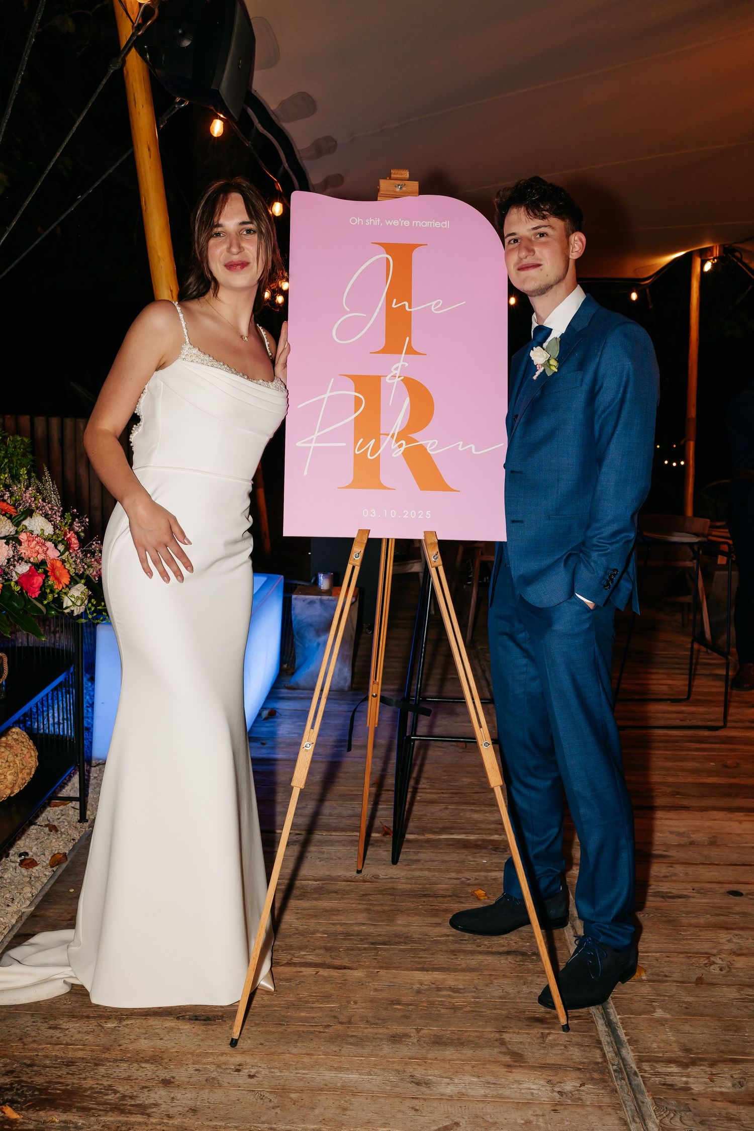 Bride and groom posing beside a pink wedding welcome sign with gold initials on an easel.