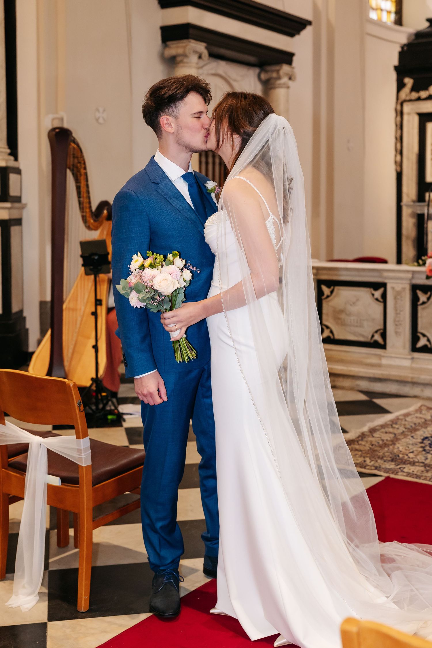 Bride and groom share romantic kiss holding bouquet before ornate church altar.