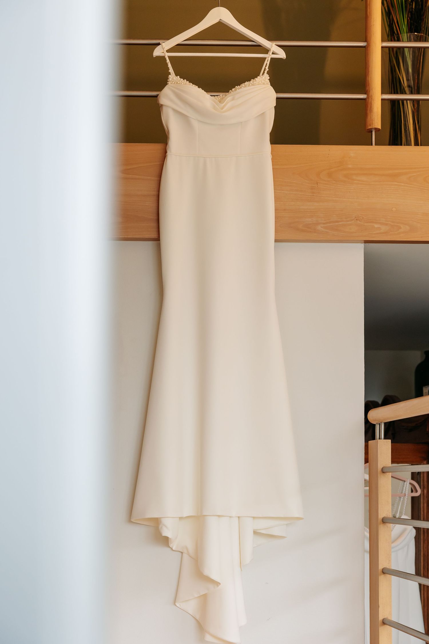 Elegant white mermaid wedding dress hanging on a wooden hanger in a bright room.
