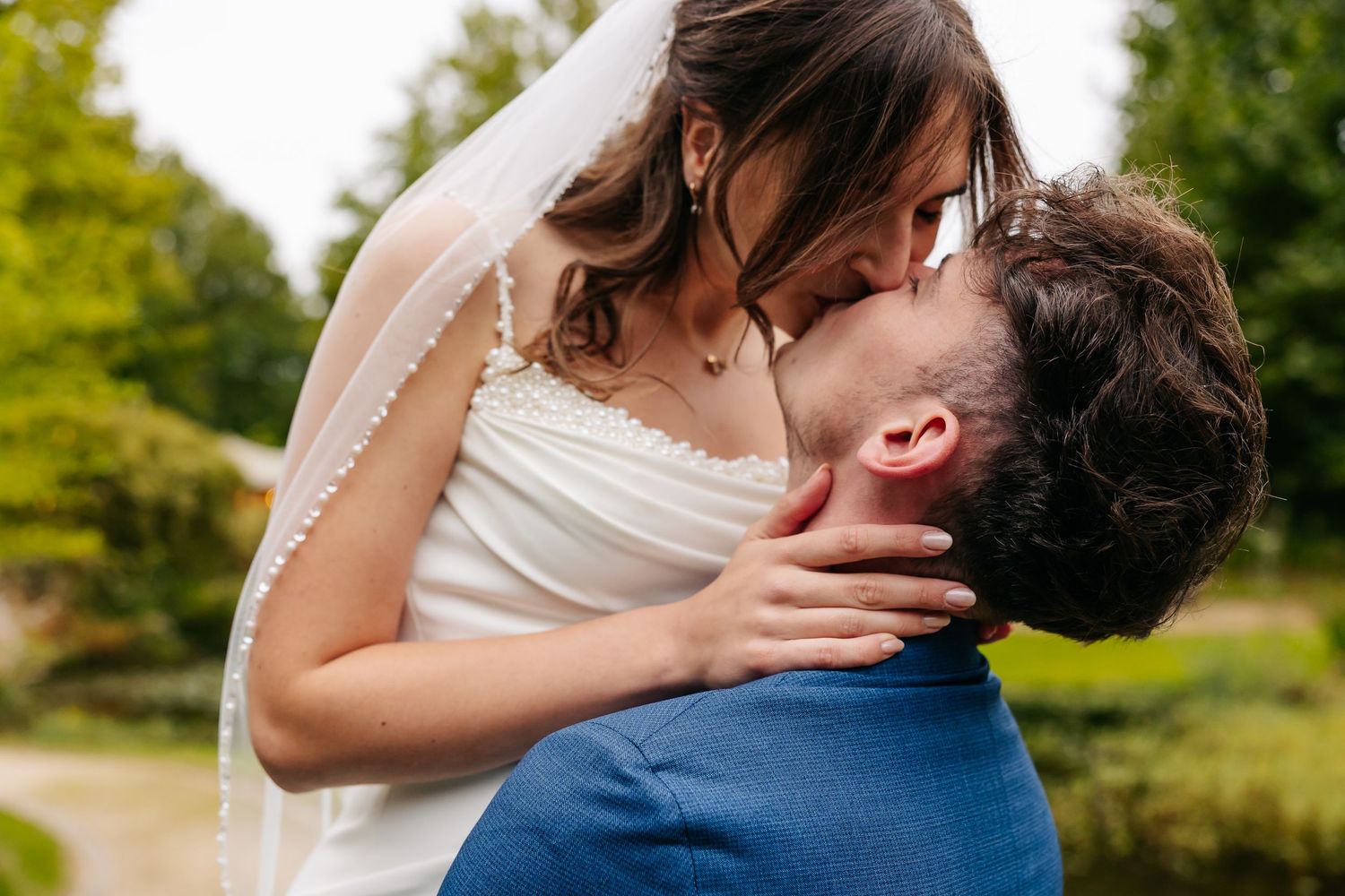 Bride and groom sharing a tender kiss outdoors surrounded by lush green trees.