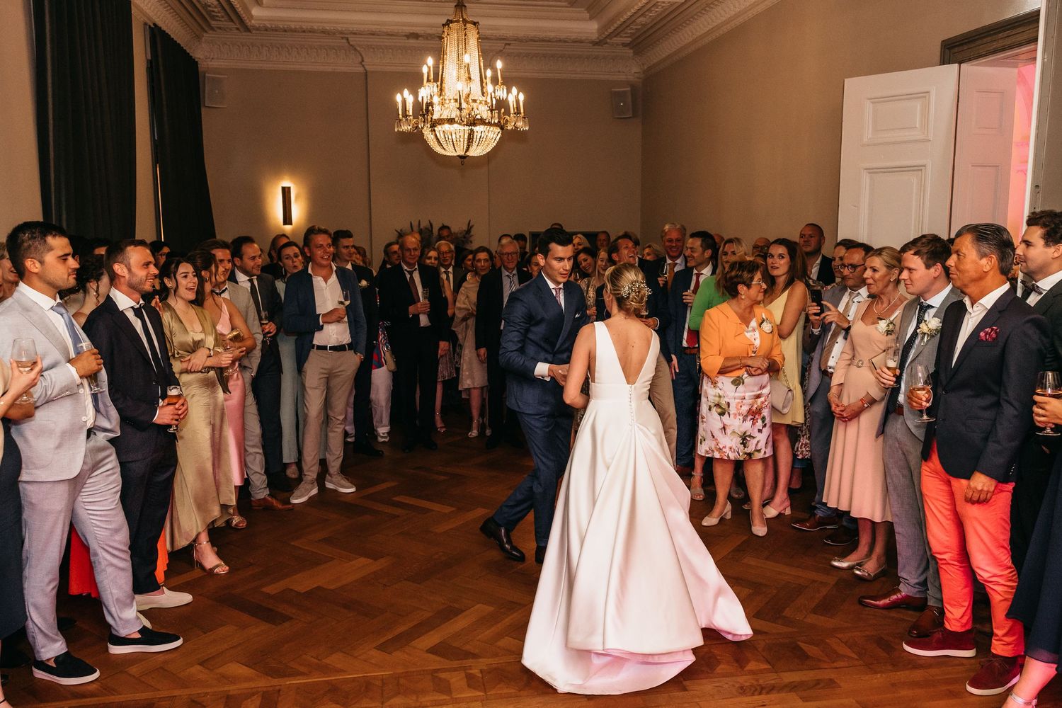 Bride and groom dance together as wedding guests clap and celebrate in chandelier-lit ballroom.