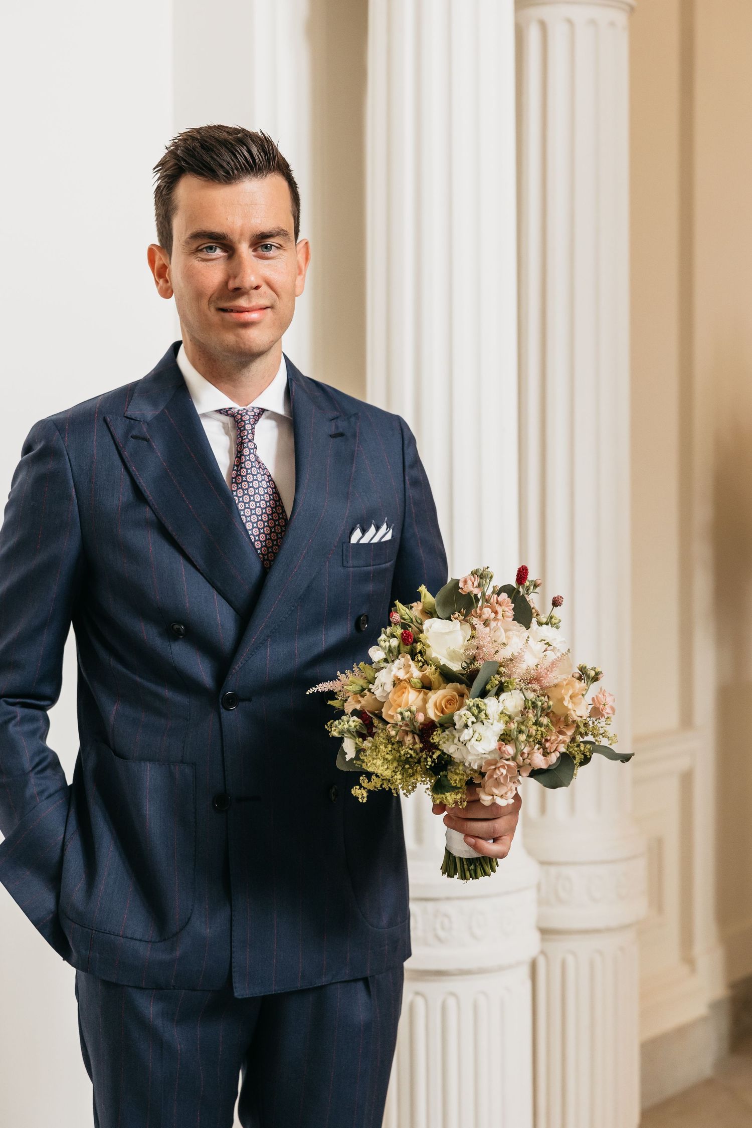 Handsome groom in navy suit smiling while holding elegant bridal bouquet near white pillars.