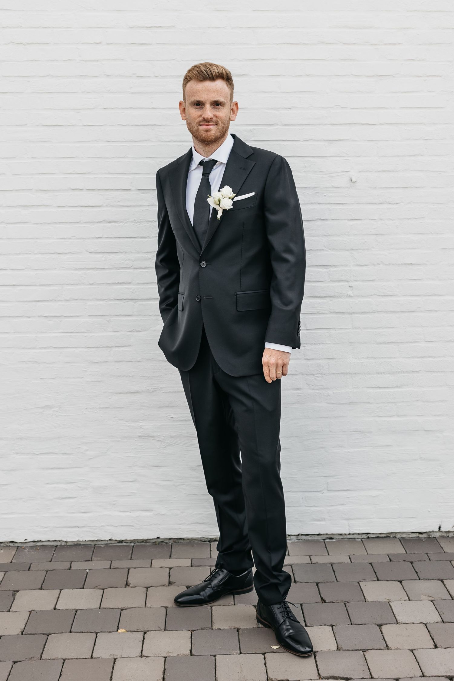 Groom in black suit with white boutonniere posing confidently against white brick wall outdoors.