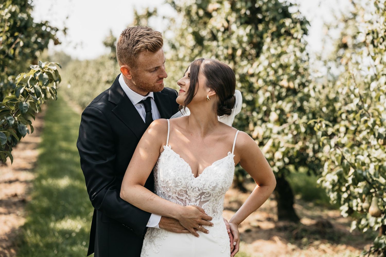 Groom tenderly embraces bride in lace wedding dress among sunlit orchard trees.
