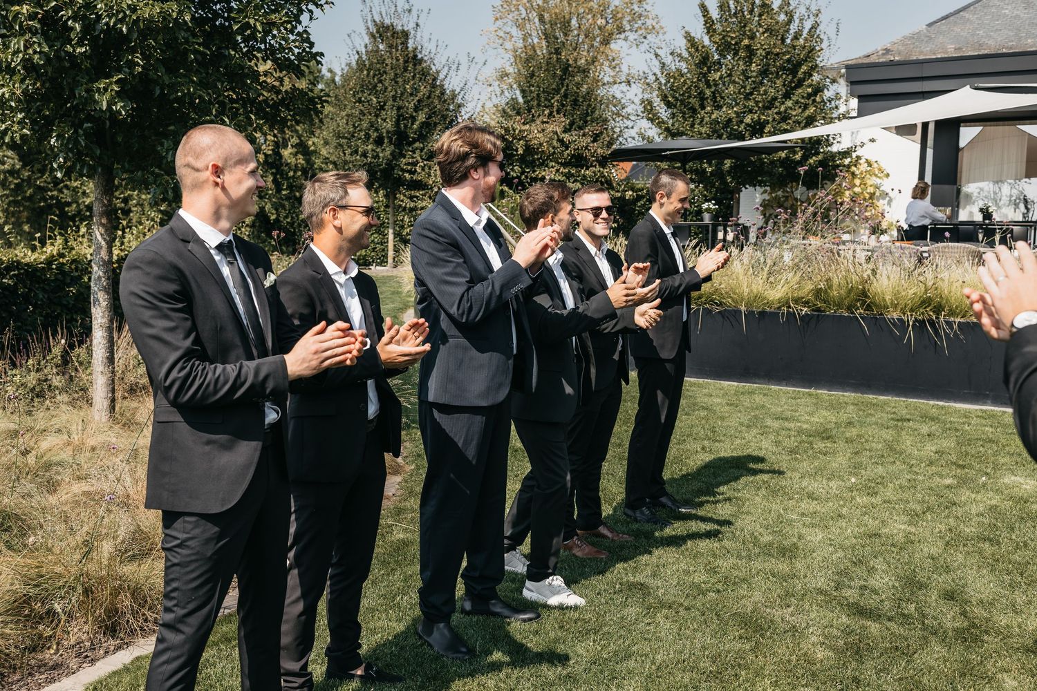 Five groomsmen in black suits clapping and smiling together on sunny green lawn at wedding.