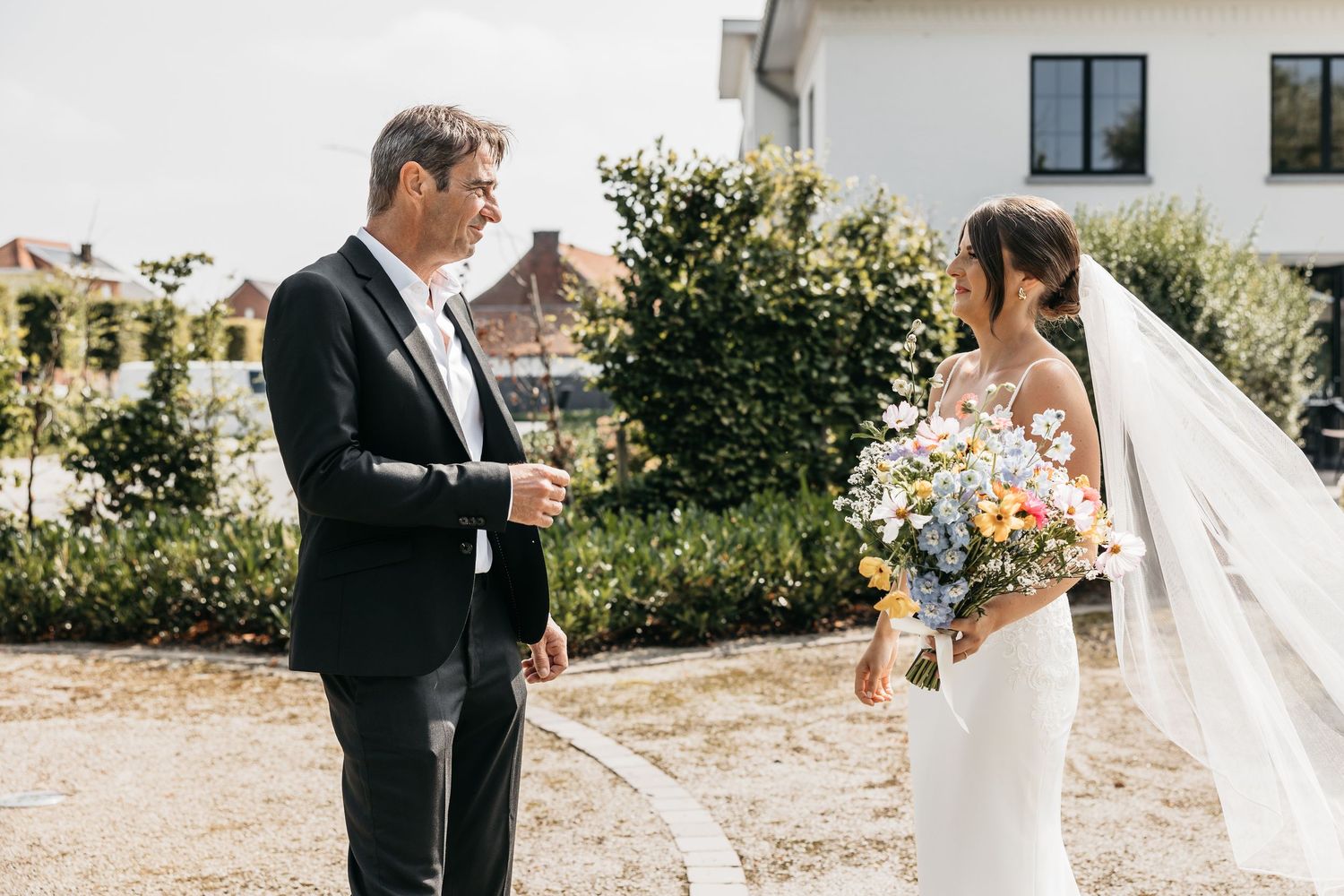 Bride in white gown with veil holds wildflower bouquet facing suited groom outdoors near hydrangeas.