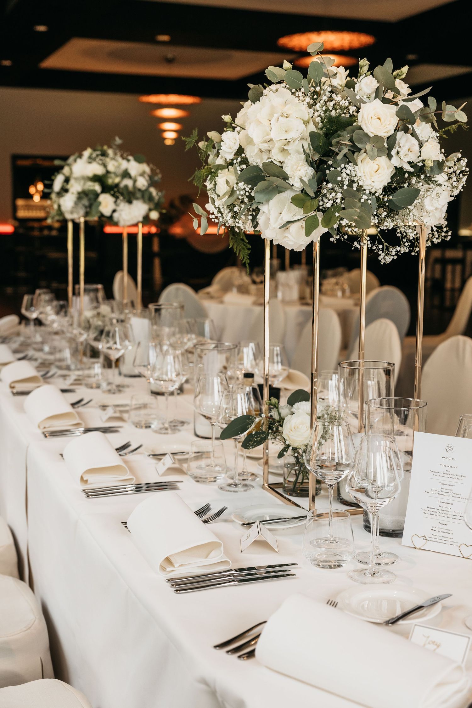 Long wedding reception table with tall gold stands holding white floral arrangements in an elegant ballroom.