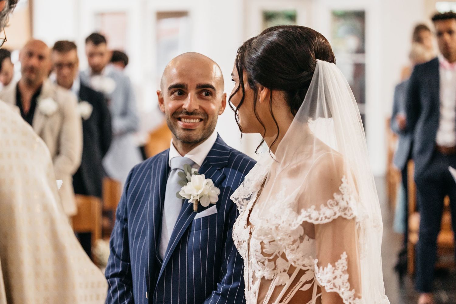 Bald groom in navy pinstripe suit smiles at bride in lace wedding dress.