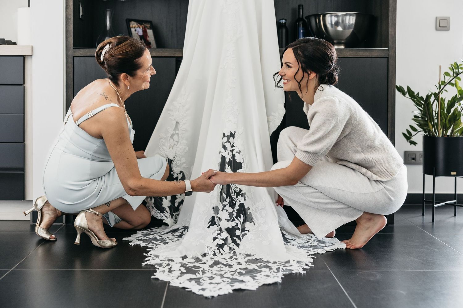 Two smiling women holding hands while crouching beside a lace wedding dress train indoors.