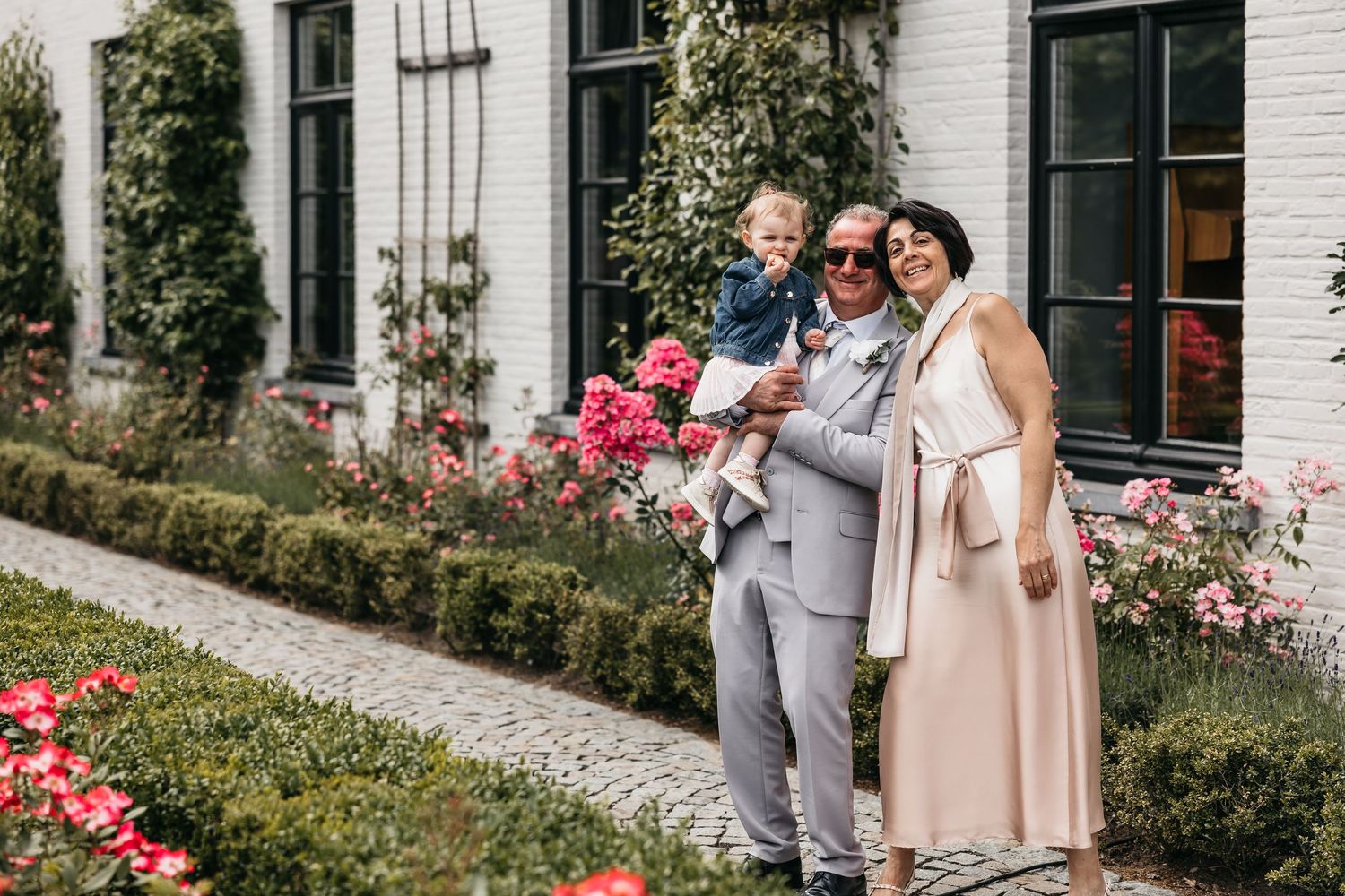 Family of three posing with young child among pink roses outside white building.