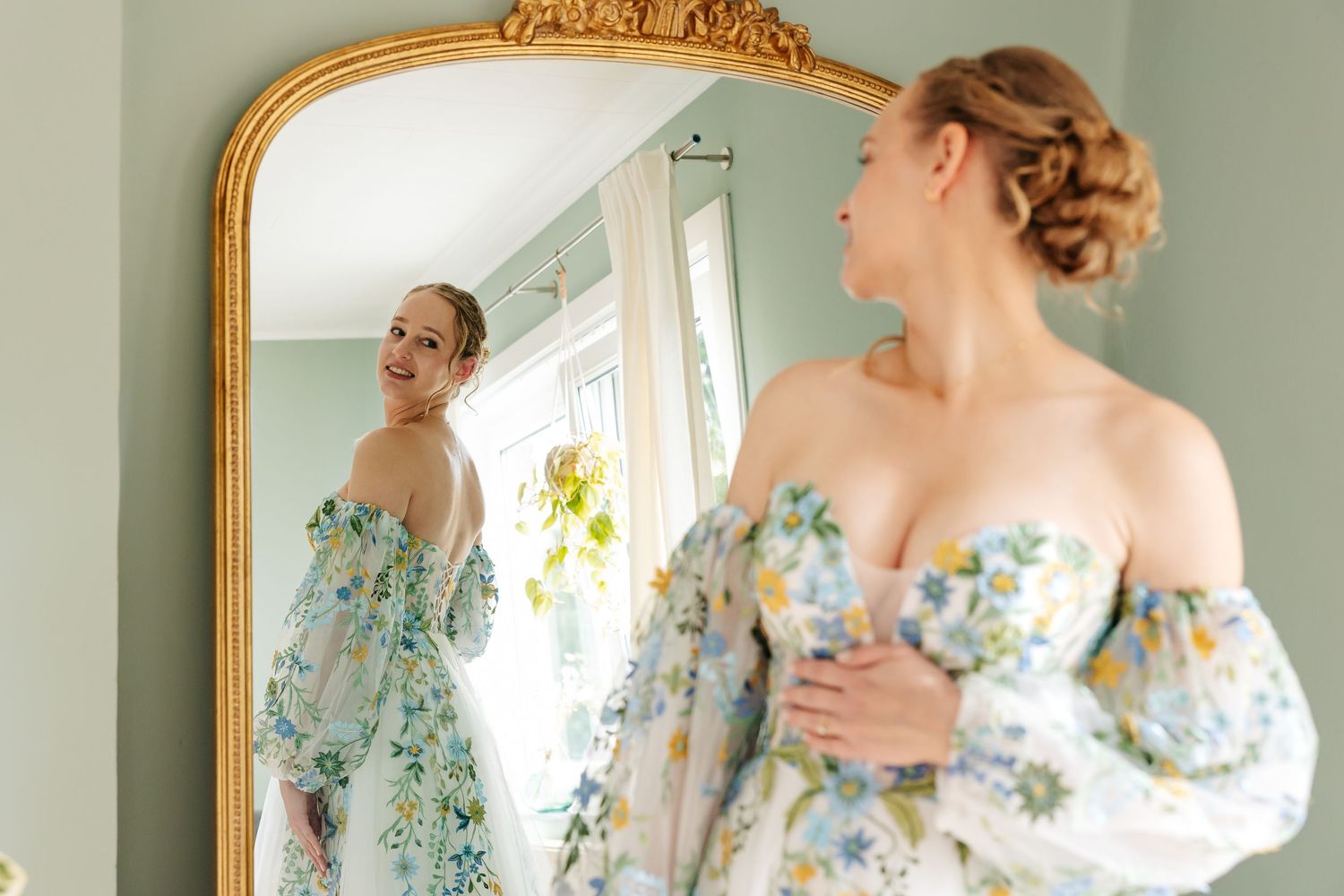 Bride in off-shoulder floral wedding dress admiring her reflection in a large gold ornate mirror.