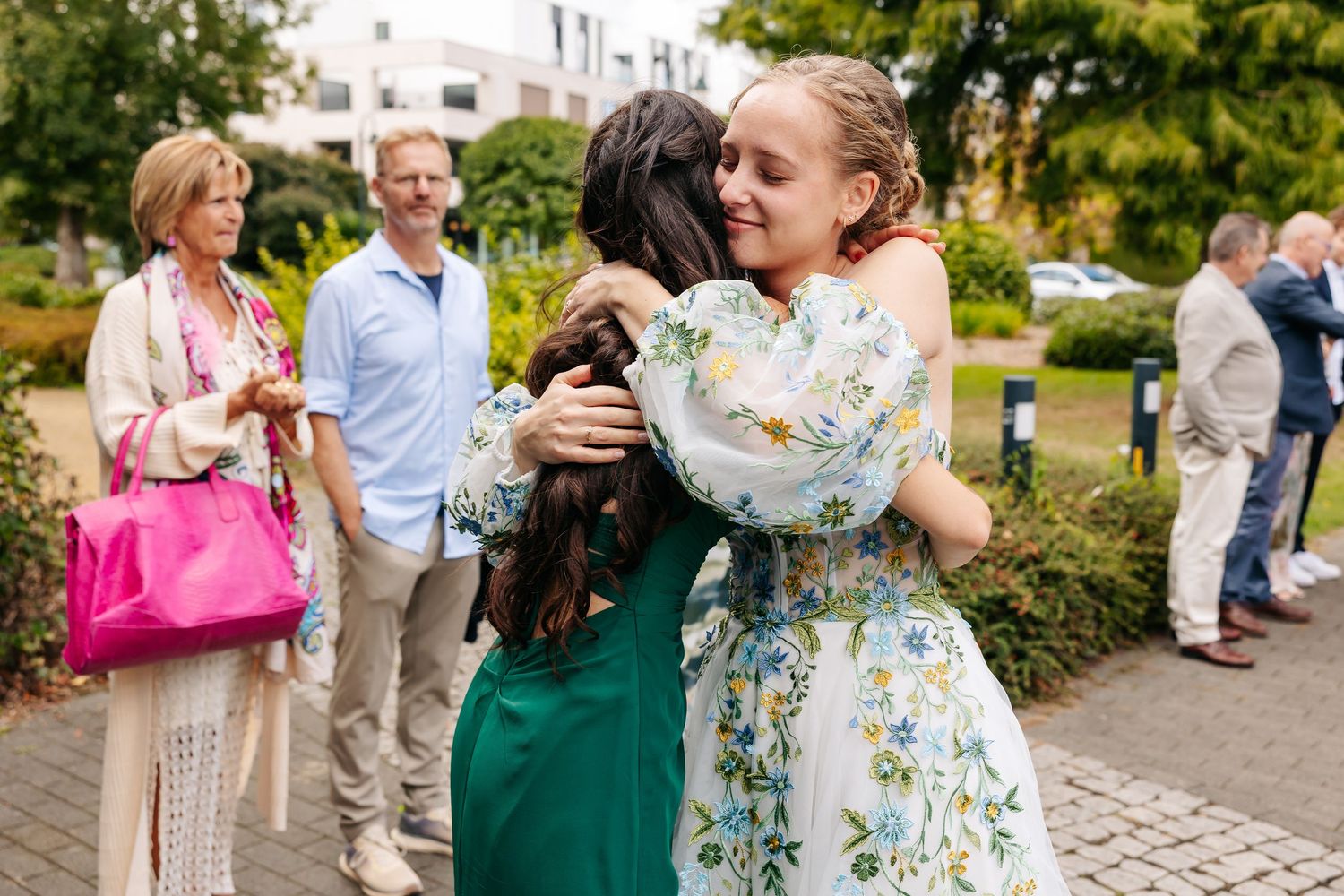 Bride in floral dress hugs a guest in green dress warmly at outdoor wedding.
