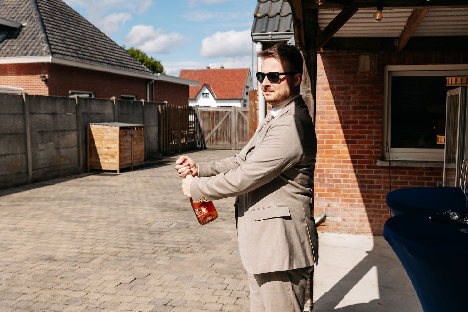 Well-dressed man in a beige suit and sunglasses holds a red item outside a brick building.