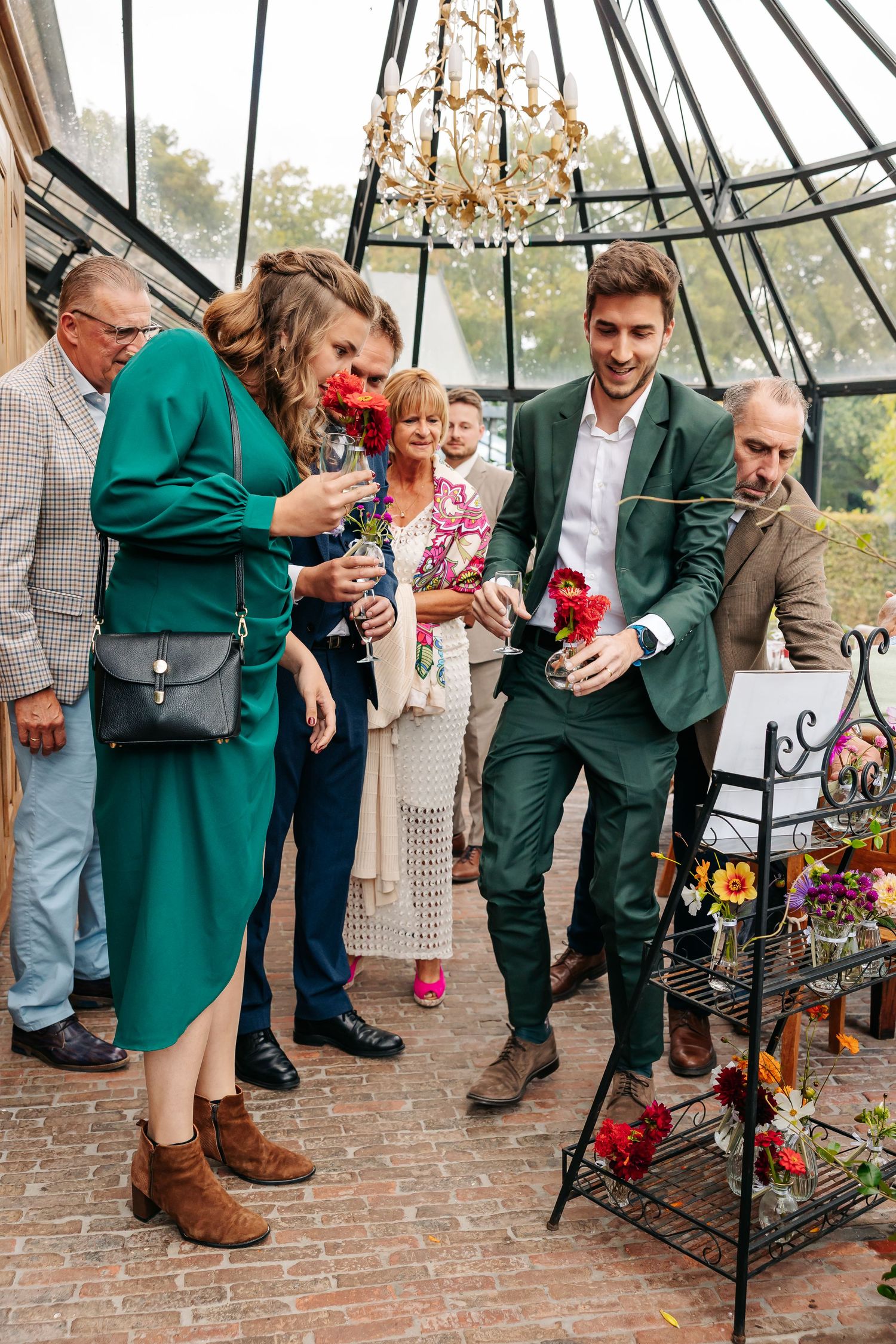 Guests in green outfits smelling red flowers at outdoor wedding floral display.