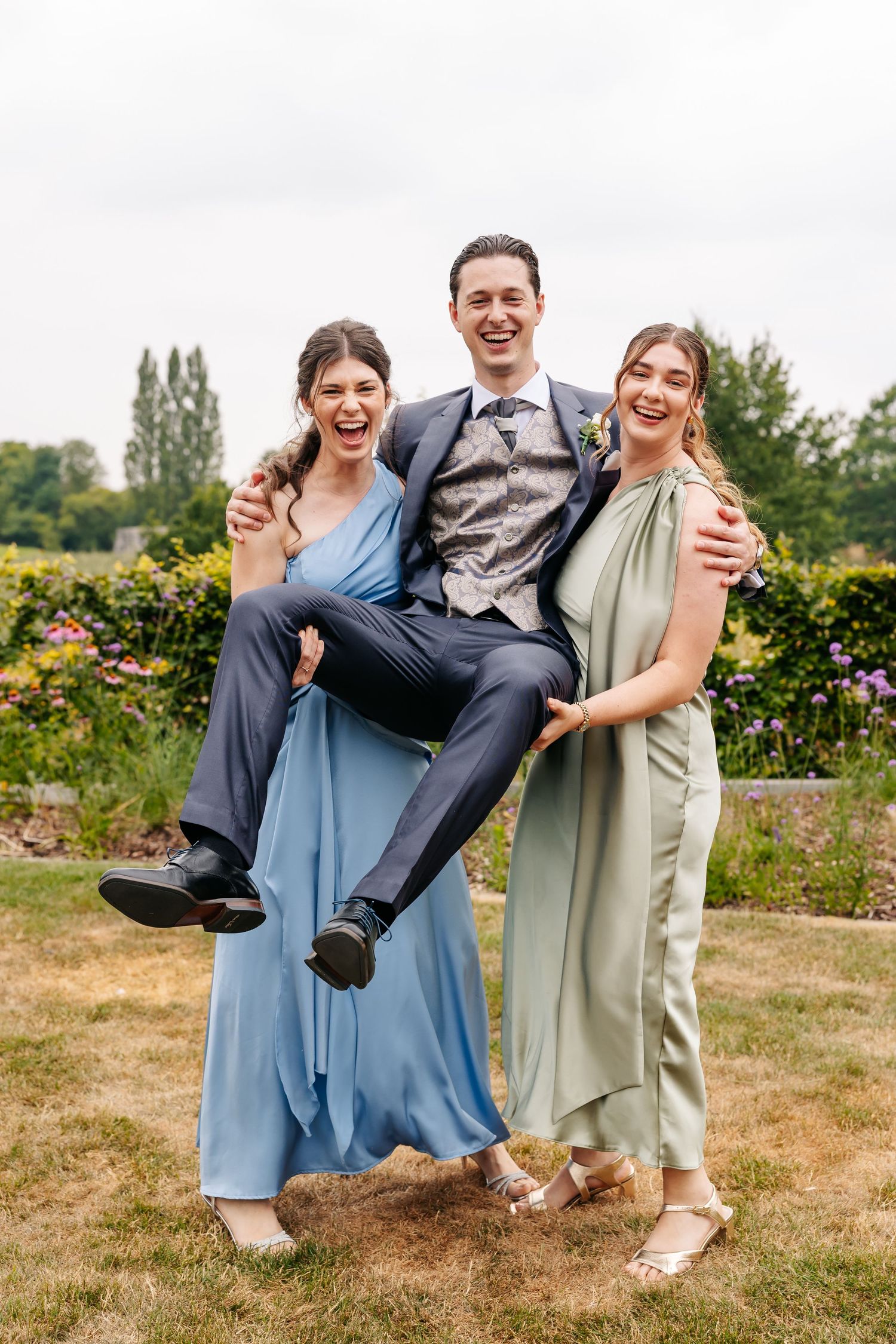 Two bridesmaids in blue and green gowns playfully lift a smiling groomsman in a garden wedding setting.