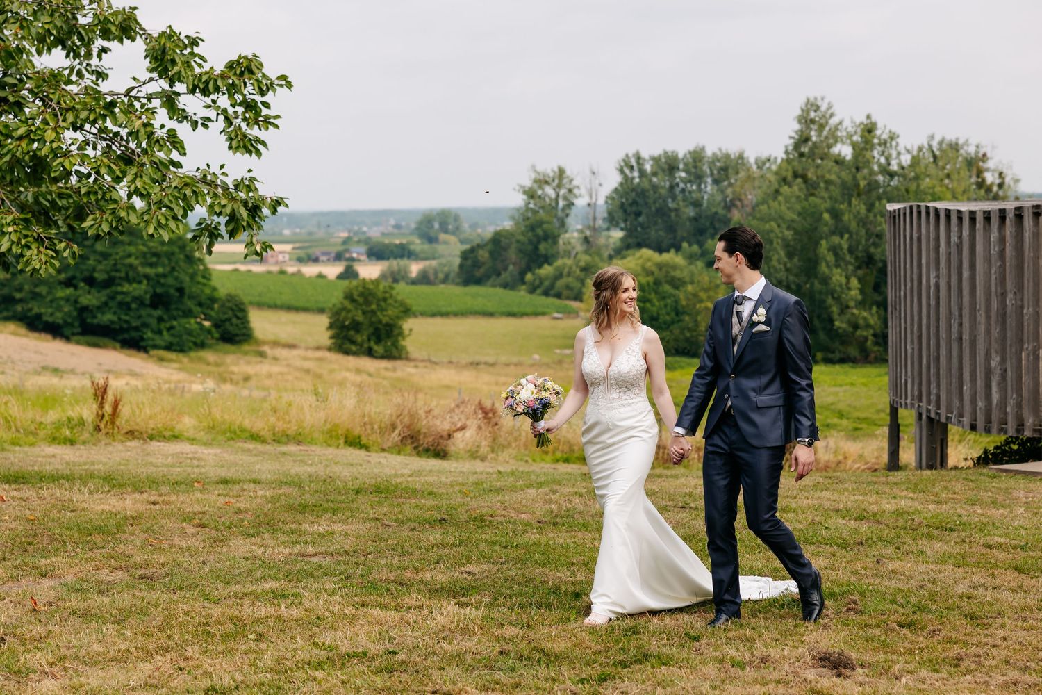 Bride and groom walking hand in hand across a sunny countryside field together.