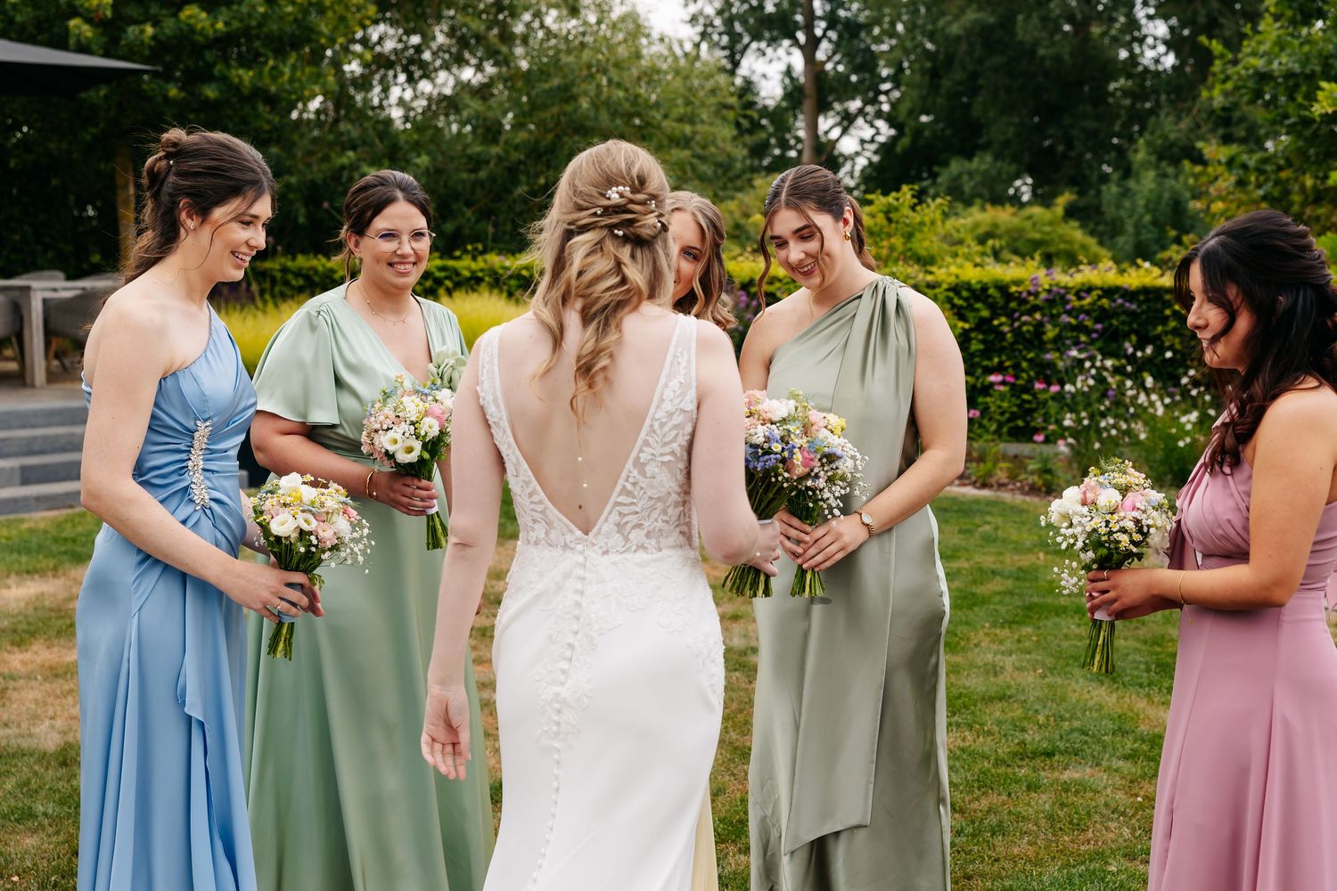 Bride in lace gown shows off open back to five bridesmaids in mismatched pastel dresses holding bouquets outdoors.