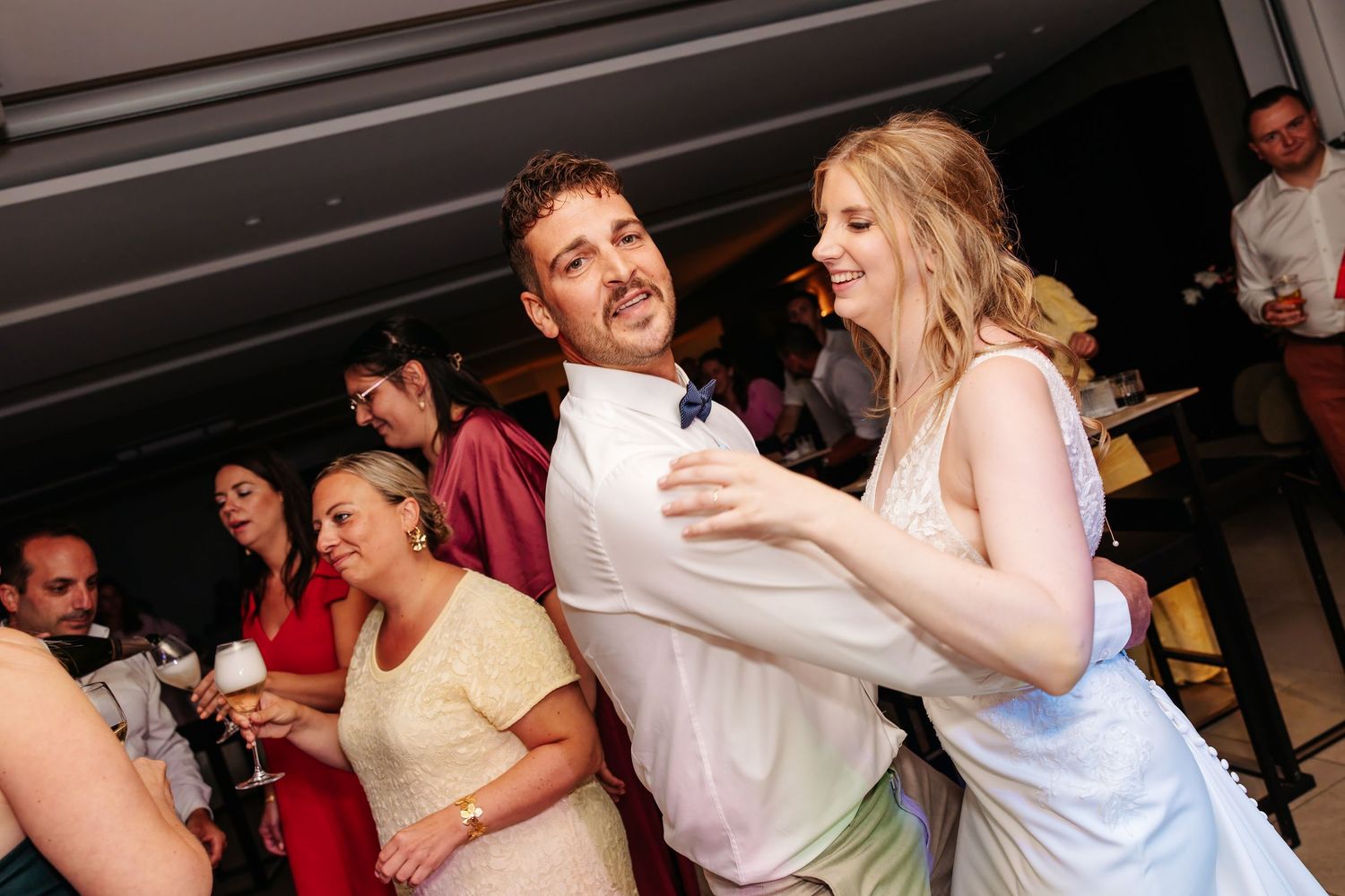 Laughing bride and groom dance together surrounded by joyful guests at wedding reception.