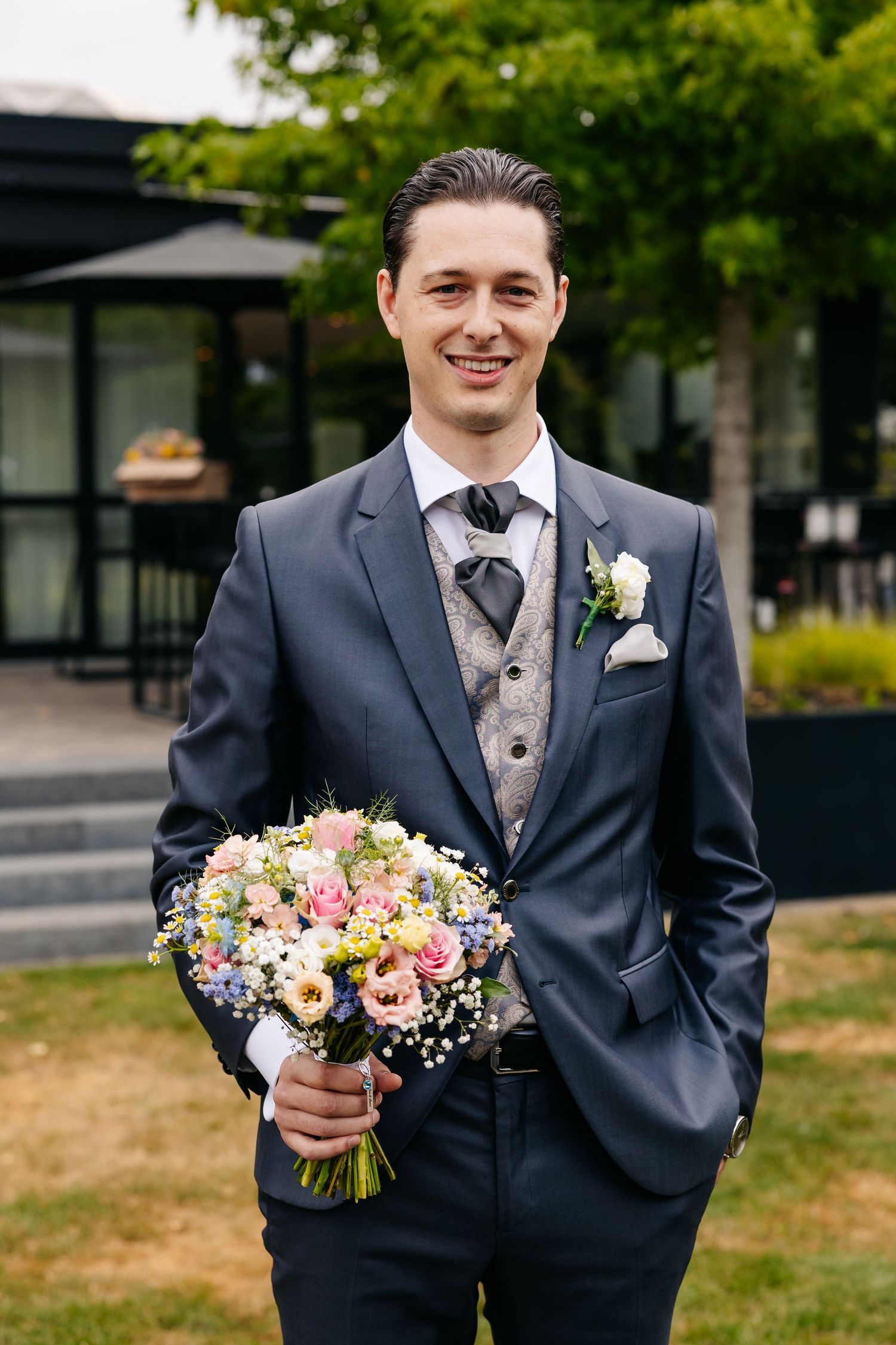 Groom in grey three-piece suit holding pink and white floral bouquet smiling outdoors.