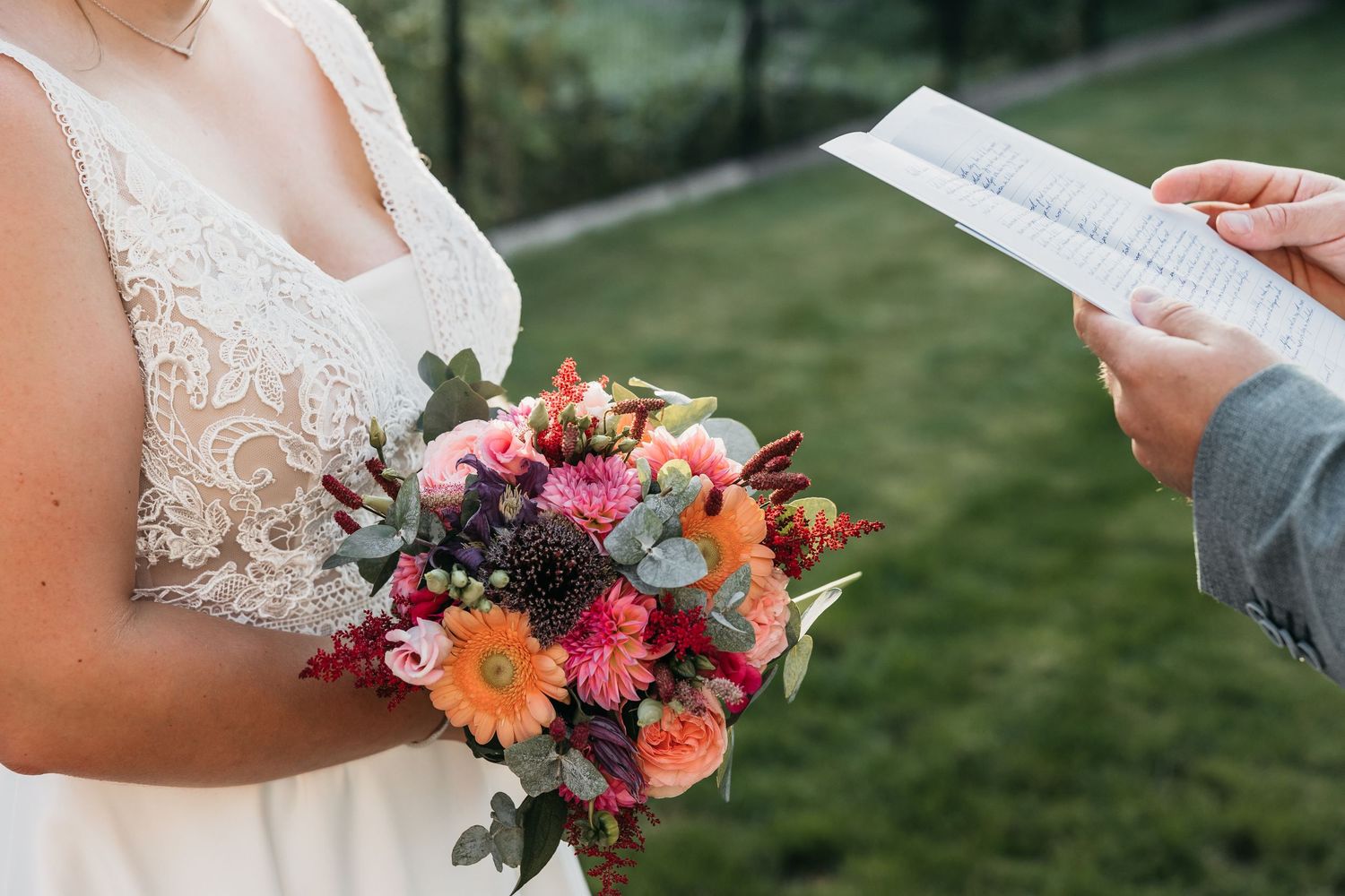 Bride in lace gown holds vibrant bouquet while groom reads wedding vows.