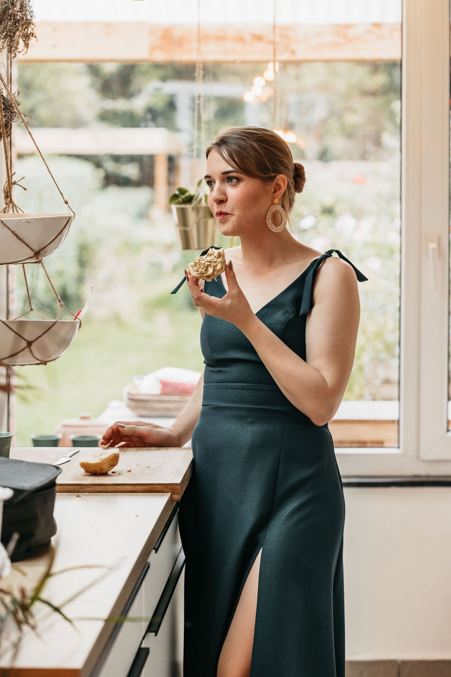 Bridesmaid in elegant dark green satin dress holding a cupcake near a window.