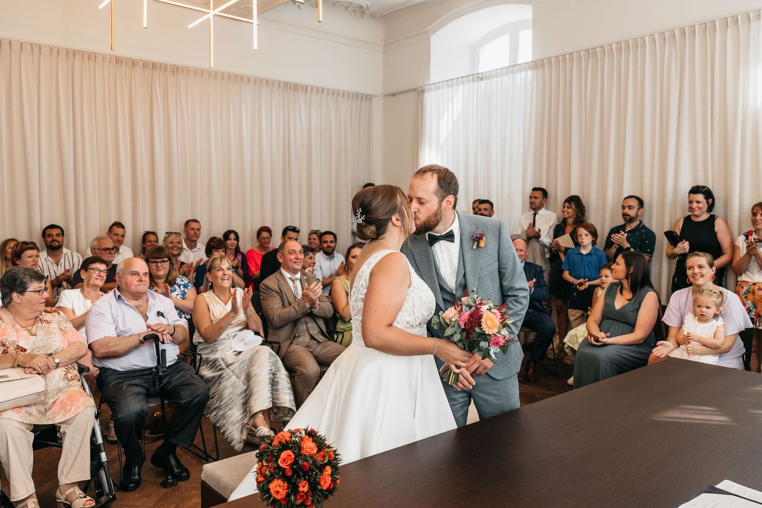 Bride and groom share first kiss at wedding ceremony while guests applaud in bright elegant venue.