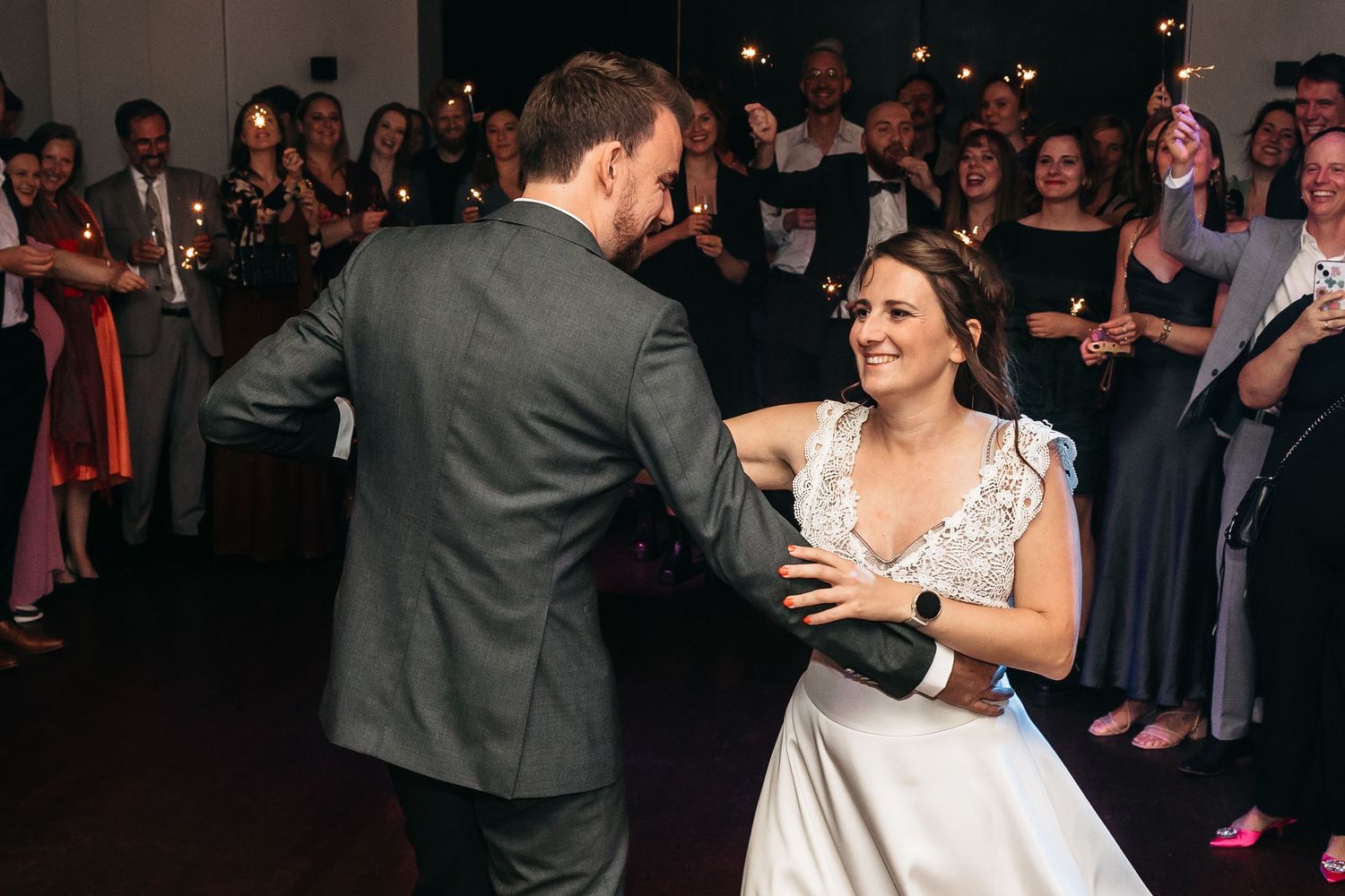 Smiling bride in lace gown dances with groom as guests hold sparklers behind them.