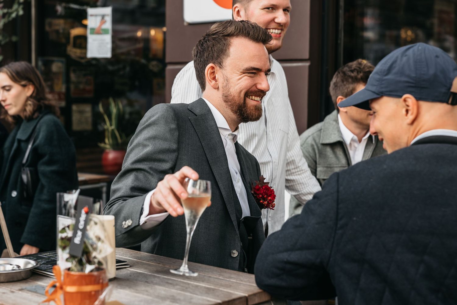 Bearded man in suit laughing while holding champagne glass at outdoor wedding reception table.