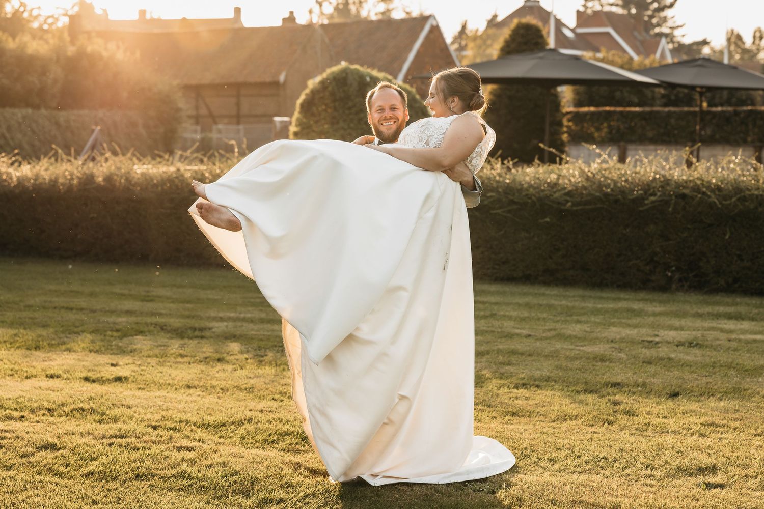 Groom lifting smiling bride in white dress outdoors during golden hour sunset.