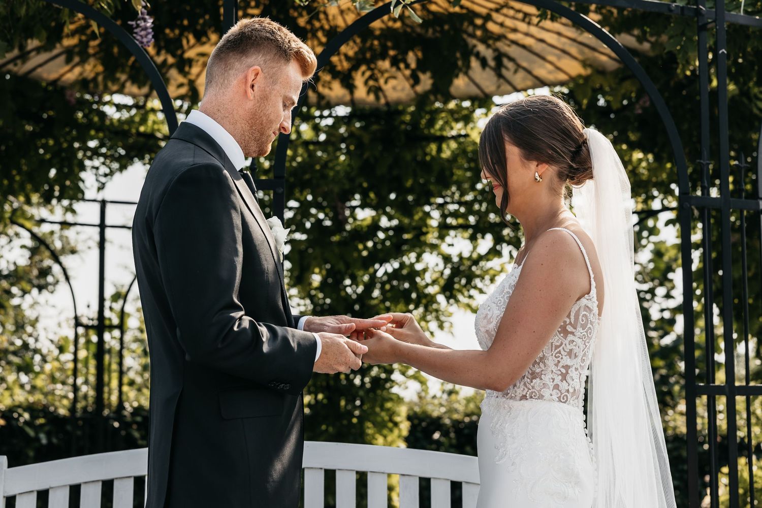 Bride and groom exchange rings tenderly under a beautiful green garden arbor outdoors.