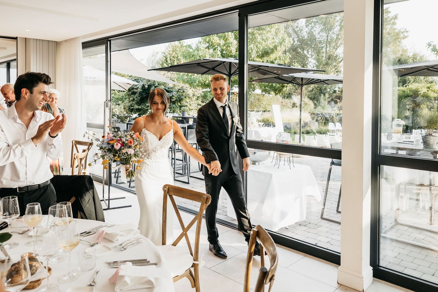Bride and groom enter bright modern wedding reception venue holding hands as guests celebrate.