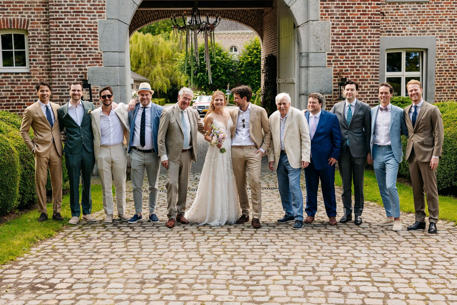 Wedding group photo of bride, groom, and groomsmen posing on cobblestone courtyard at elegant estate.