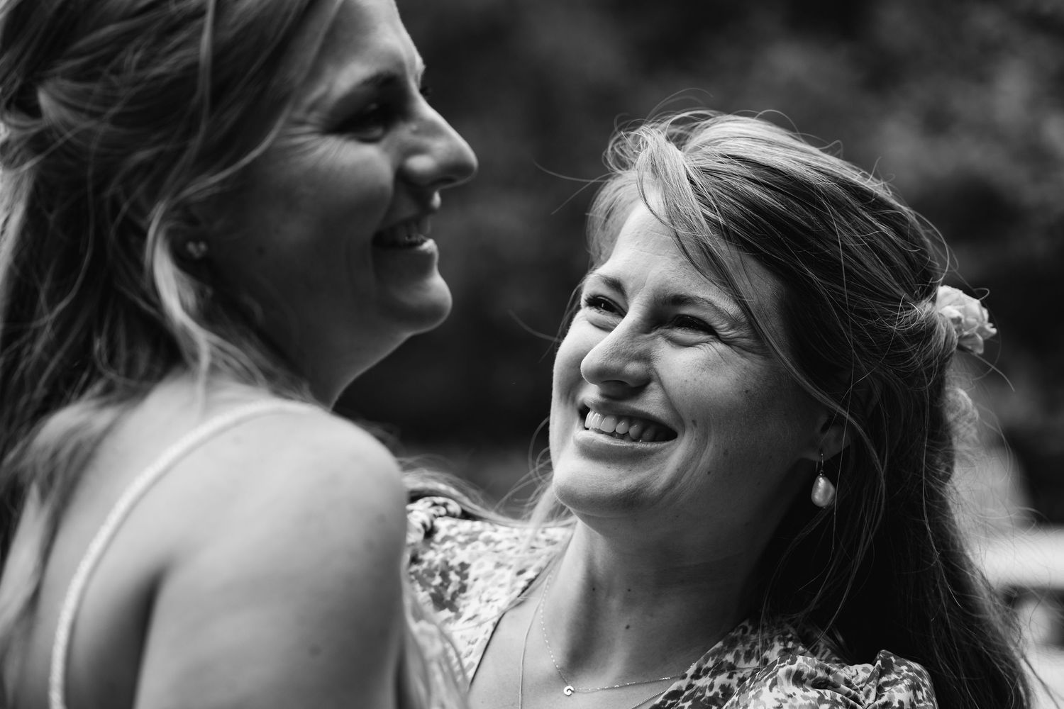 Black and white close-up of two women smiling and laughing joyfully at an outdoor wedding.