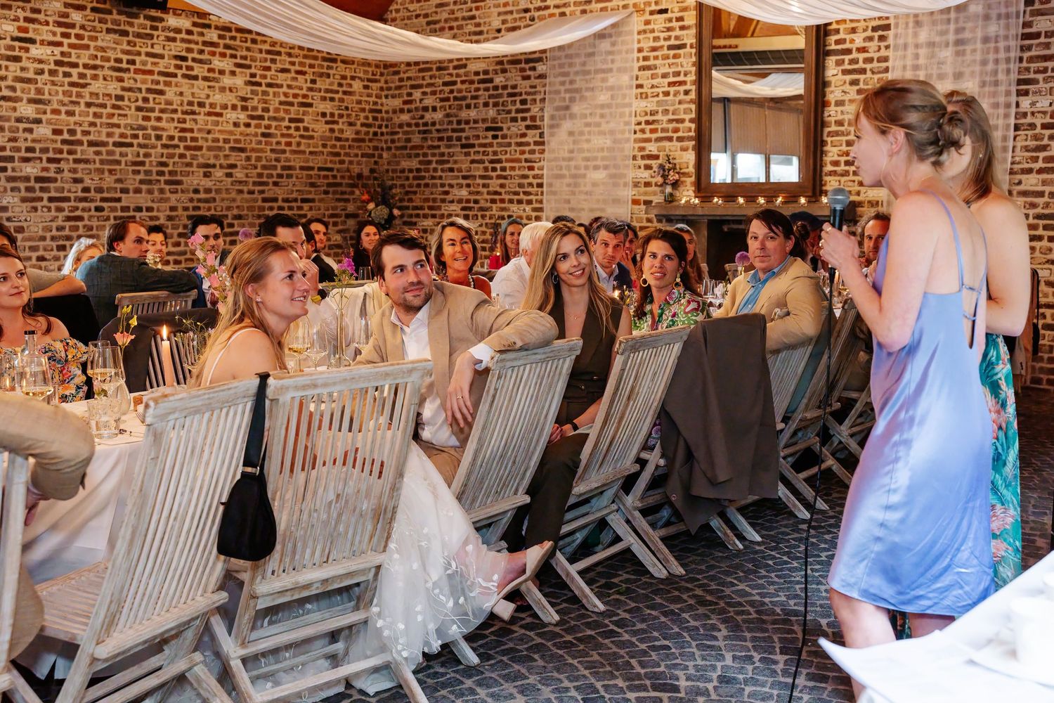 Wedding guests seated in wooden chairs inside a rustic brick venue, smiling at a speaker during the reception.
