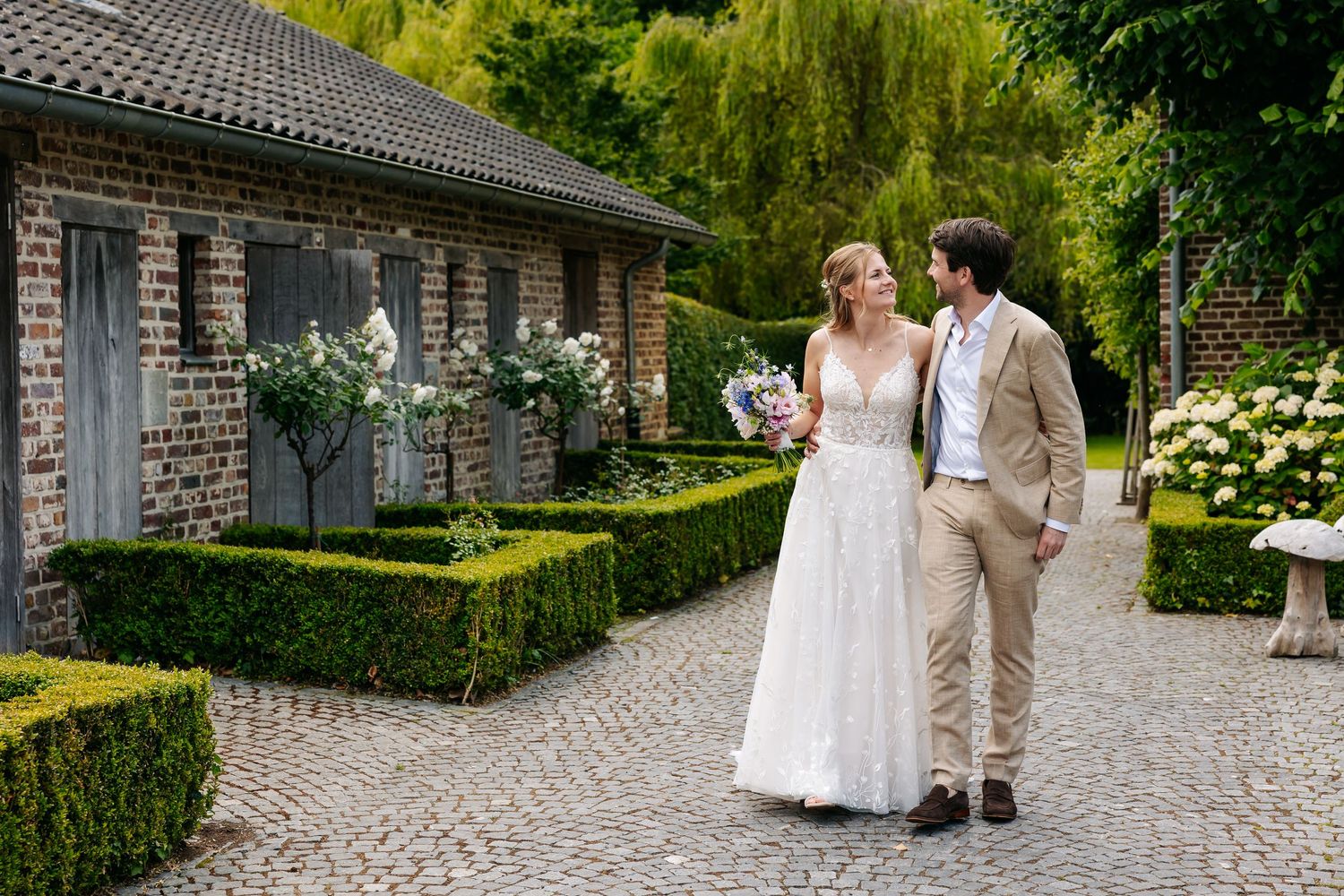 Bride and groom walking hand in hand through formal garden with boxwood hedges at estate.