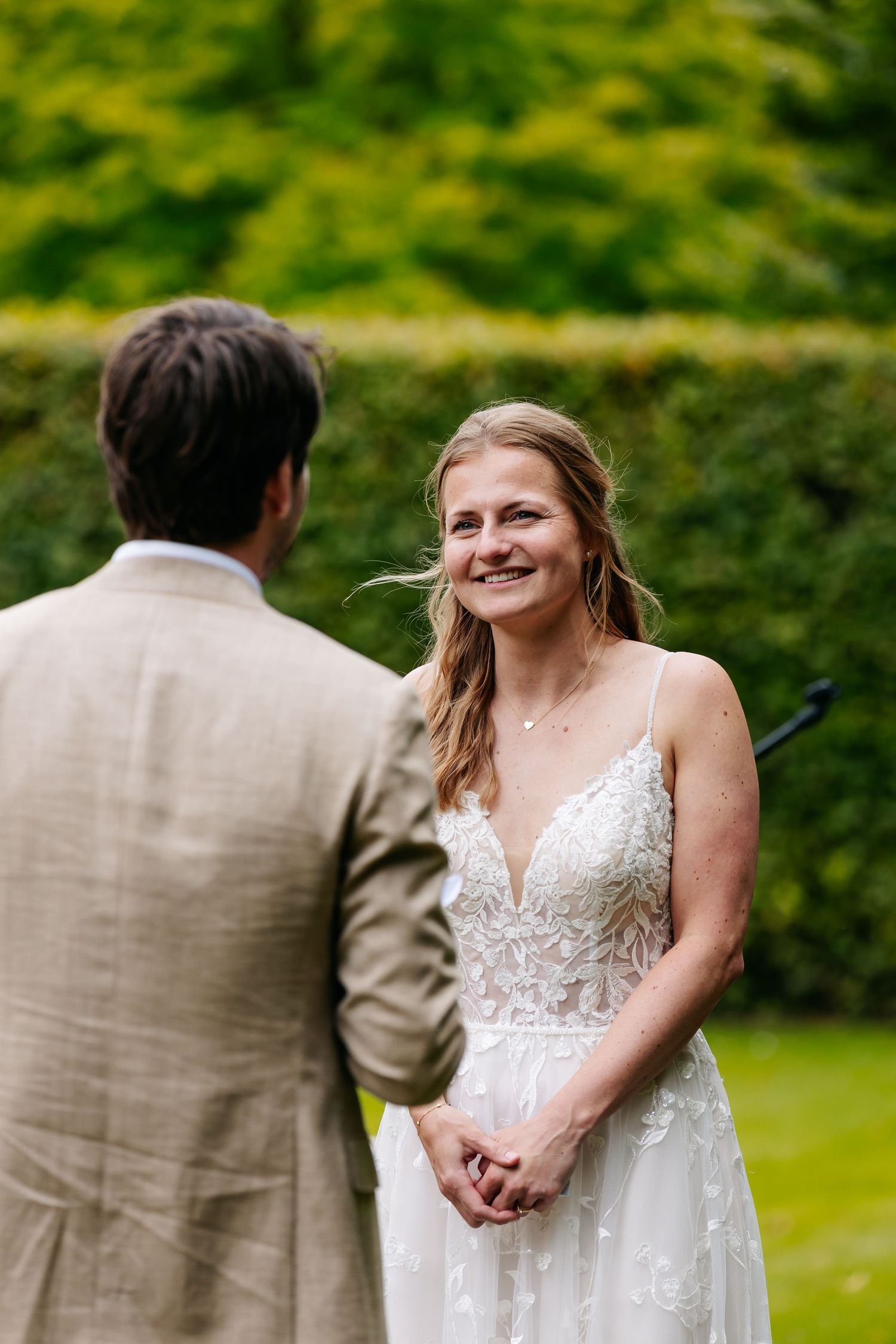 Smiling bride in beige lace gown holds grooms hands during heartfelt outdoor wedding ceremony.