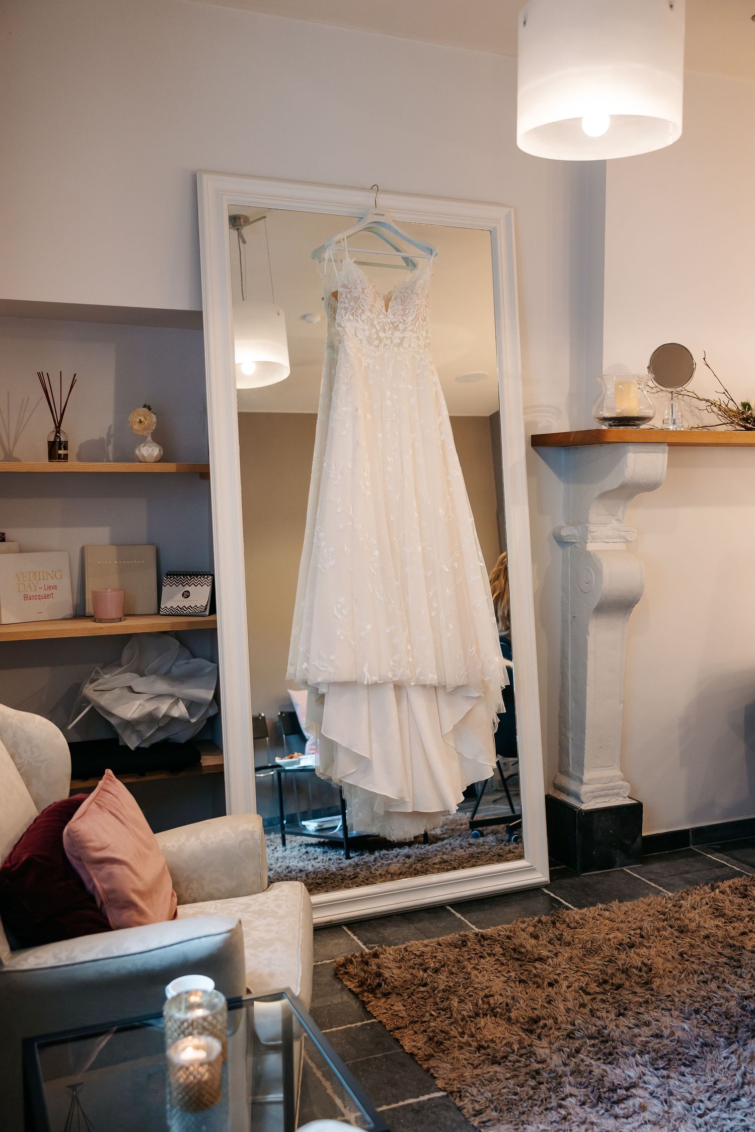 White lace wedding dress hanging on a mirror in an elegant bridal boutique fitting room with cozy decor.