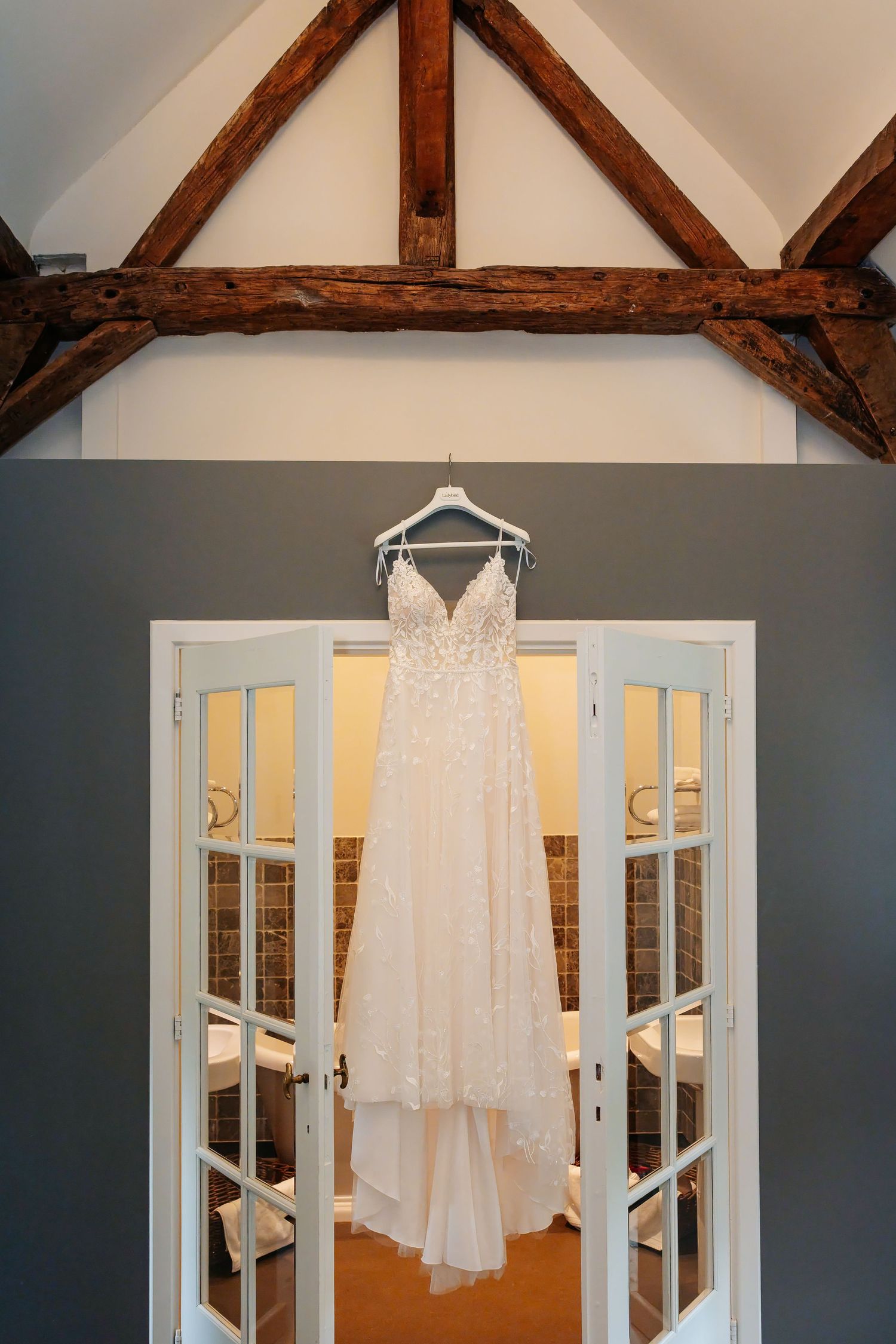 Elegant lace wedding dress hanging over French doors with rustic wooden beam ceiling in bridal suite.