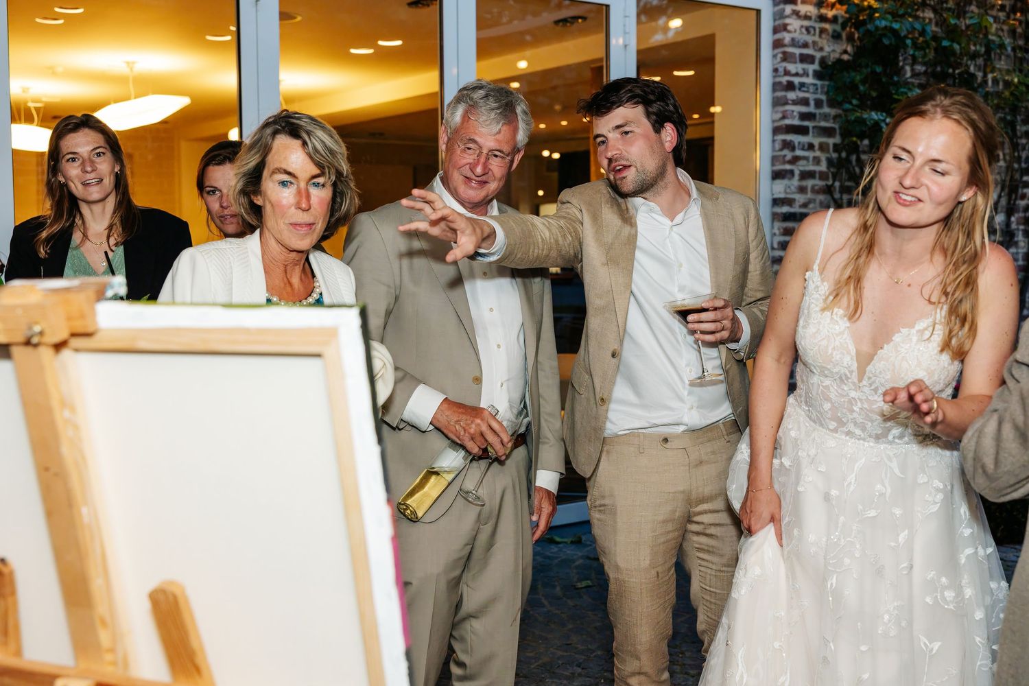 Guests and bride admiring live wedding painting on easel at indoor reception venue.